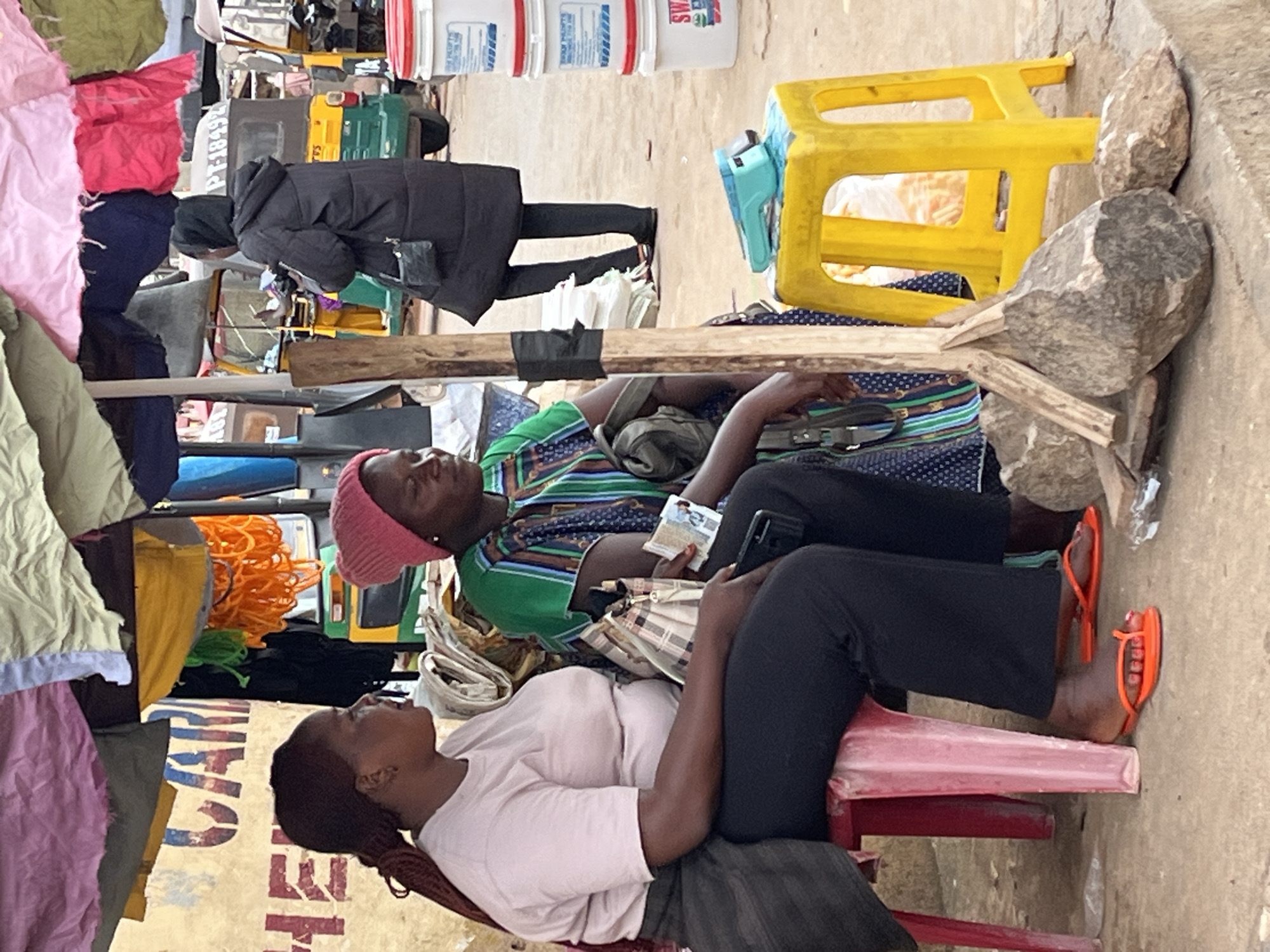 The image depicts two young women resting outdoors in what appears to be a market or roadside stall. The setting is cluttered with various goods, including bolts of fabric in different colors, coiled orange material, and what look like water containers or buckets stacked in the background. A bright yellow plastic stool is visible, along with large rocks that seem to be part of the environment.
The two women are seated on red plastic stools. The woman on the left is wearing a green and blue patterned top and a pink headscarf. She is leaning back with her eyes closed, holding a few banknotes and a mobile phone. The woman on the right is wearing a light pink t-shirt and black trousers. She is also leaning back, looking upwards with her eyes closed. She has orange flip-flops on her feet. Both women appear to be resting or napping.
The ground is a dirt or dusty surface, and the overall impression is one of a warm, possibly sunny day given the outdoor setting and the subjects' attire. The clothing of the women and the vibrant colors of the goods suggest a lively, perhaps tropical or sub-tropical, environment.
In the background, some white buckets with red and blue labels are visible. A partial view of some text is present on a banner or sign behind the woman on the right, reading "CA" and "TI" in large letters. The overall scene conveys a sense of everyday life and a moment of respite amidst a busy environment.