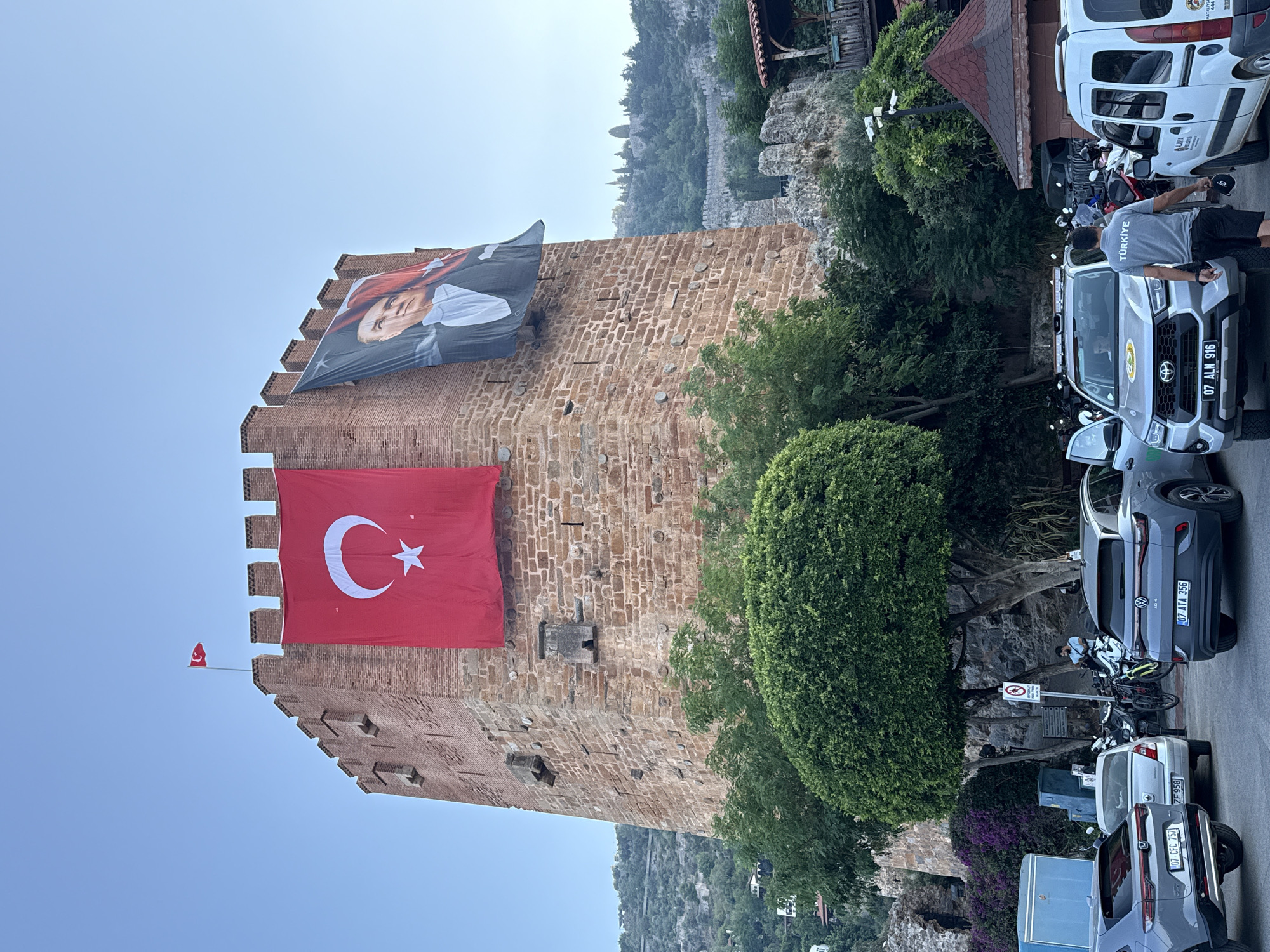 In Alanya, Turkey, a large brick tower, likely a historical defense structure, is prominently featured. A Turkish flag, red with a white star and crescent, hangs from the upper section of the tower. To the right of the flag, a large banner displays a portrait of a man in a suit. Green trees and foliage surround the base of the tower, with a clear blue sky overhead. In the foreground, several vehicles are parked, including a gray SUV with a license plate reading "07 ATA 356", a white van, and other cars. A man wearing a gray t-shirt with the word "TÜRKIYE" and black shorts is walking away from the camera. The background shows a hillside covered in trees and a fortified wall, suggesting the tower is part of a larger ancient complex.