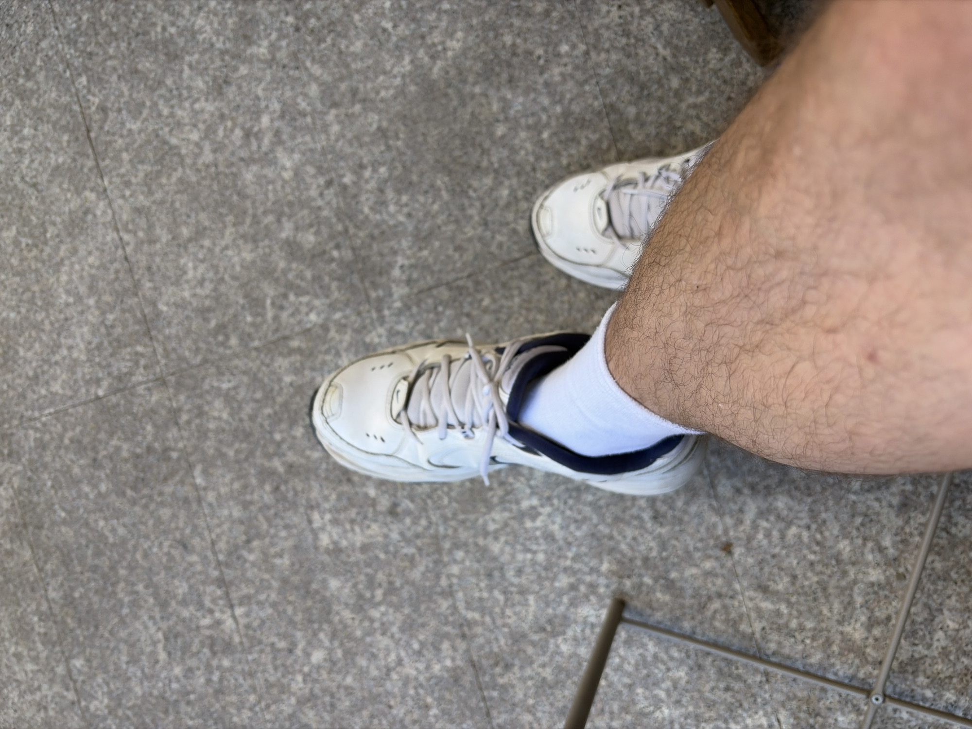 Overhead shot of two white sneakers, one partially obscured by a right human leg covered in hair and wearing a white sock. The shoes are on a gray, stone-like floor. The lower-right of the shot includes metal chair parts. No location details present.