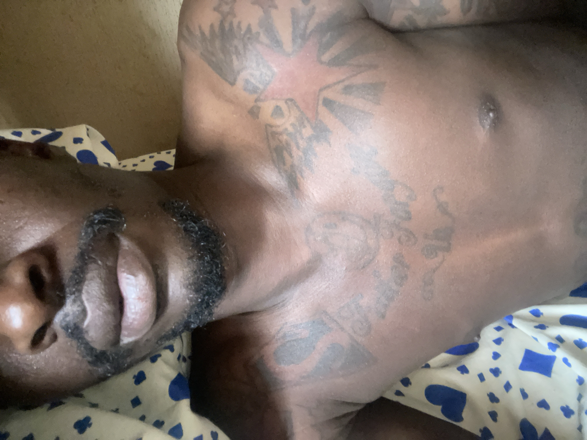 A man with dark skin is lying on a bed with a patterned blue and white sheet. He is shirtless, and visible tattoos are present on his upper torso and shoulder. The man has a beard and mustache. The image appears to be taken in a dimly lit indoor setting. The location is not identifiable as Jos, Nigeria from the visual information provided.