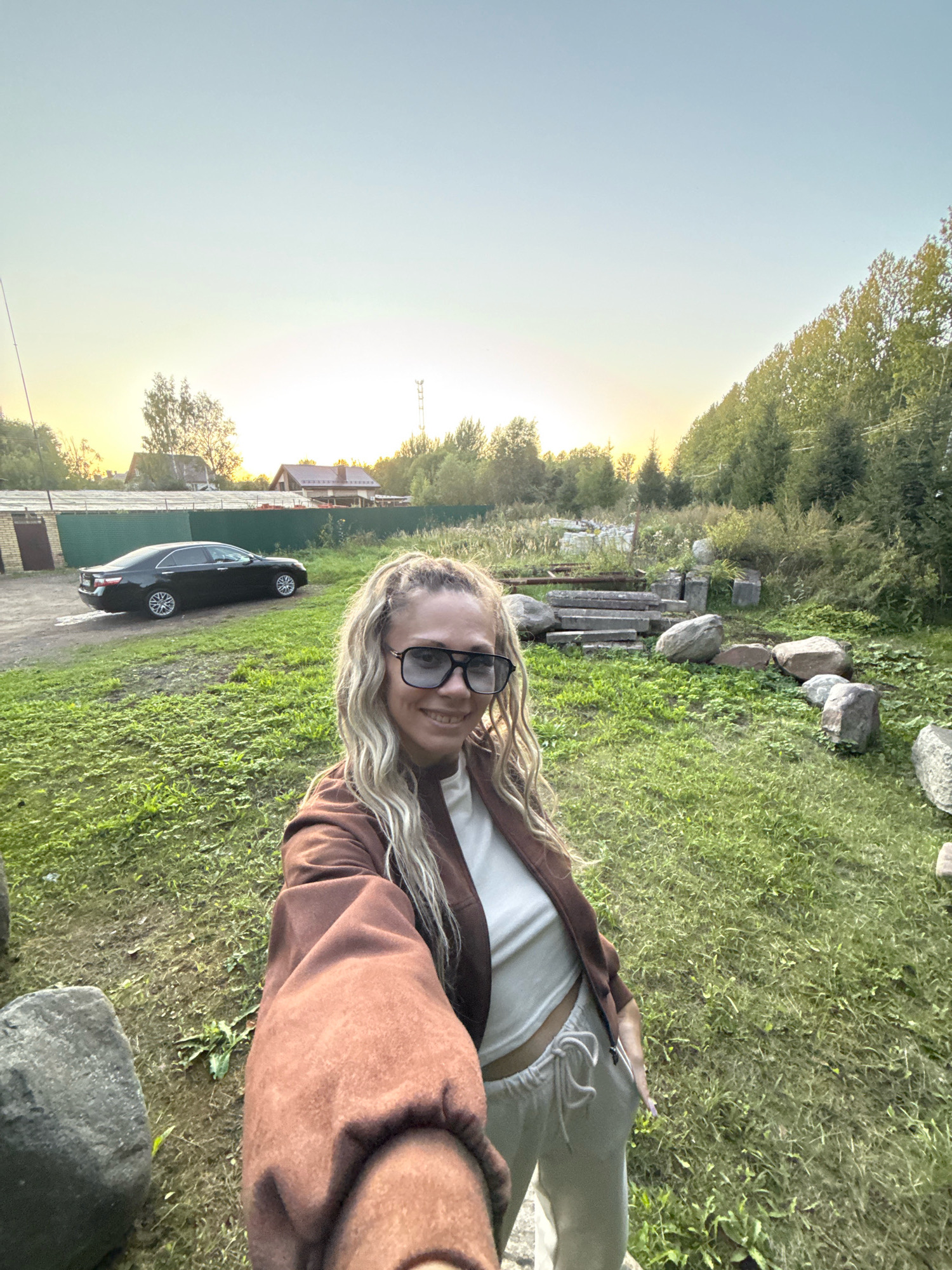 A woman with blonde dreadlocks and sunglasses is taking a selfie outdoors at sunset. She is wearing a brown jacket over a white tank top and light-colored sweatpants. In the background, a black sedan is parked on a gravel driveway next to a green fence. Behind the fence are two houses. The landscape is grassy with scattered large rocks and trees. The sky is a pale yellow and blue, indicating the sun is setting. The location appears to be a rural area near Rusanovo, Russia.