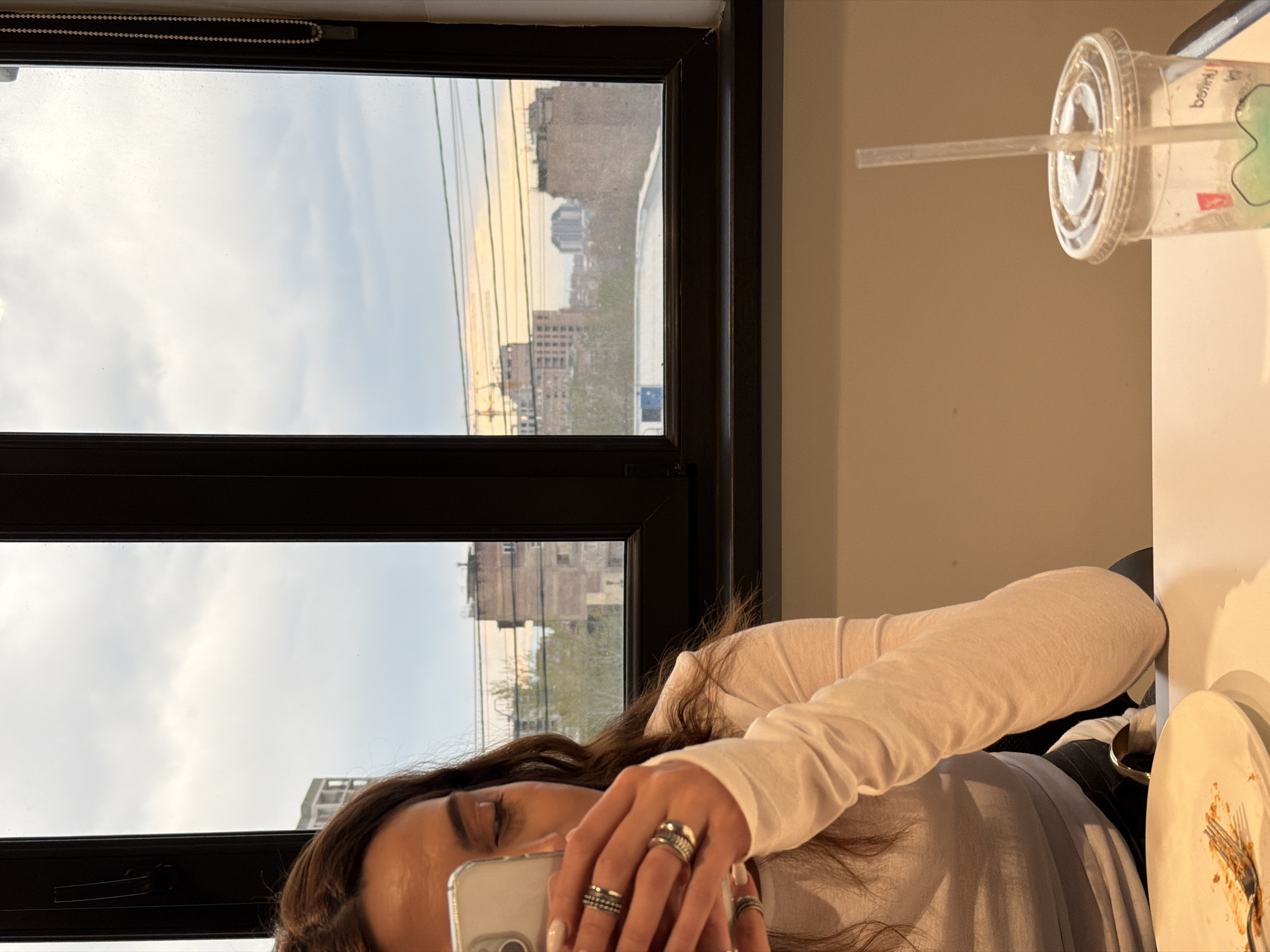 A person with long brown hair and wearing a long-sleeved white shirt is taking a photo with a smartphone. The person has multiple rings on their fingers. Next to the person, on a white surface, is a clear plastic cup with a lid and straw, and a used plate with food remnants. Behind the person, a dark window frame reveals a cityscape under a cloudy sky, with buildings visible in the distance. Based on the architectural style and location, the city is identified as Yerevan, Armenia.