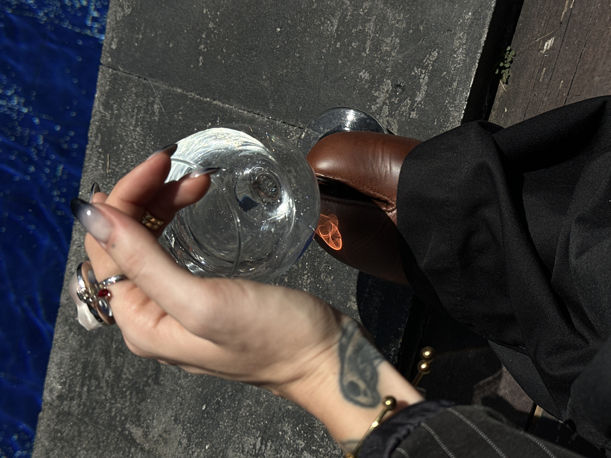 A person is holding a wine glass filled with a clear liquid. The person has dark, ombre-styled fingernails and is wearing multiple rings on their fingers, including one with a red stone. A tattoo is visible on the person's wrist. They are wearing black trousers and brown leather shoes. The scene appears to be outdoors, with a stone surface and a body of blue water in the background. The lighting suggests it is daytime. The location is identified as North Kuta, Indonesia.