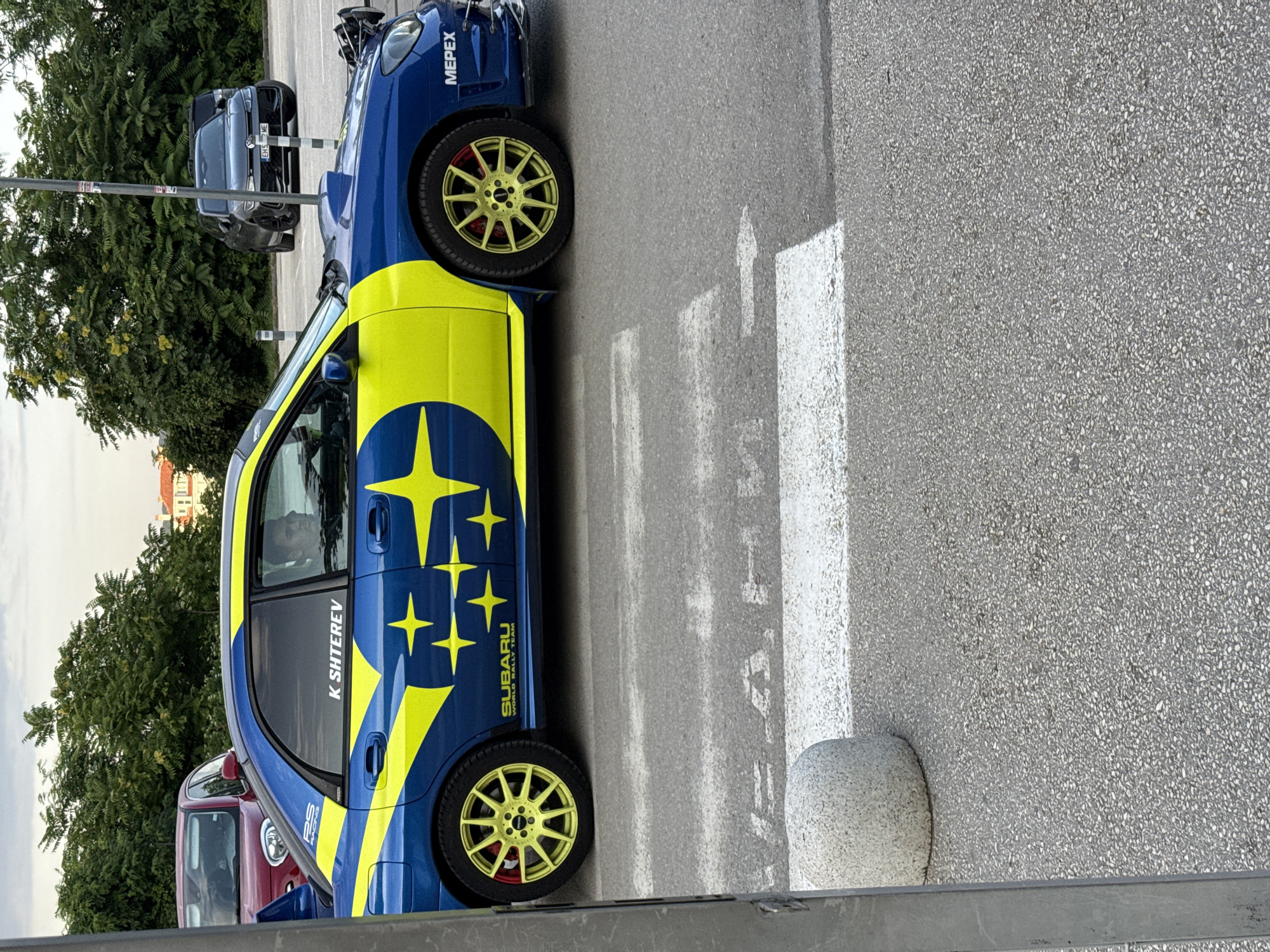 The image shows a blue and yellow Subaru WRX STI parked on a street. The car has a racing livery with the Subaru World Rally Team logo and several yellow stars. The text "K SHITEREV" is visible on the driver's side window. The car is equipped with gold aftermarket wheels and red brake calipers.
Behind the Subaru, a dark gray hatchback is parked, and a red car is partially visible to the left. A row of trees with green foliage lines the left side of the frame. The background shows a building and a cloudy sky, suggesting it might be daytime with overcast weather. The car is positioned on a paved surface with white pedestrian crossing markings. A round concrete bollard is visible near the right edge of the image.
The scene appears to be a street or parking area, likely in a European city given the location context of Pleven, Bulgaria. There are no people clearly visible in the image. The overall impression is of a performance car displayed in an urban or semi-urban environment.
