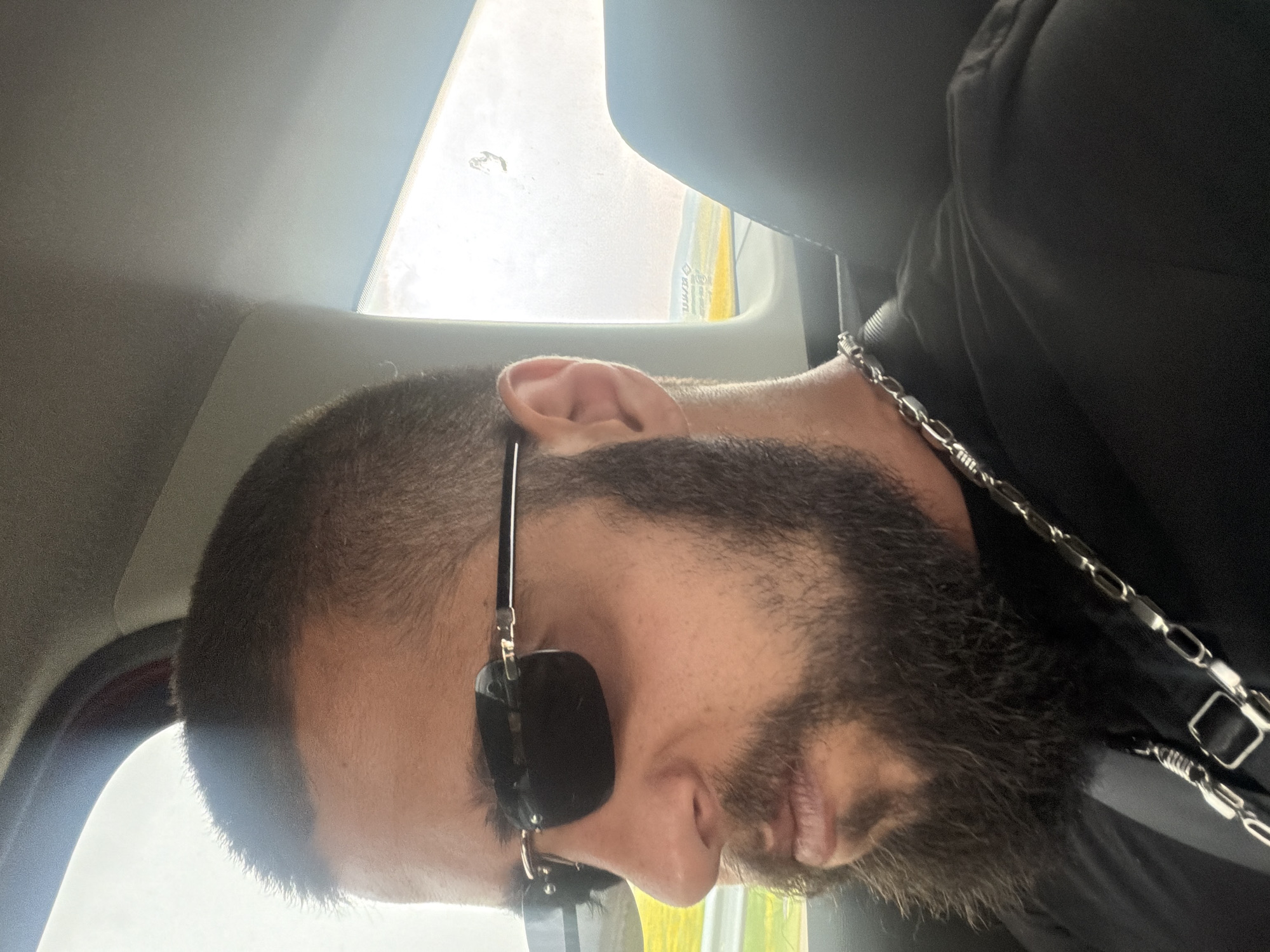 The image shows a close-up, side-profile view of a man's head and shoulders, taken from inside a vehicle. He has short, dark hair and a full, dark beard. He is wearing dark sunglasses, which obscure his eyes. A silver chain necklace is visible around his neck, draped over his dark clothing. The lighting suggests it is daytime, with bright light coming from a window, possibly indicating a sunny or overcast sky. The background is blurred but appears to be a car interior with a window showing a hint of the outside world. There is no visible text in the image. The man's expression is neutral, and the overall mood is casual. The setting is likely a car traveling in or around Omarchevo, Bulgaria.