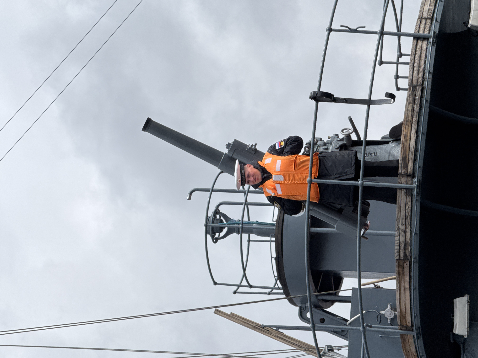 A man in a navy uniform and orange life vest stands next to a large gun on a ship. The gun is mounted on a platform with railings. The sky is overcast. The man is wearing a white hat. The ship appears to be old, with wooden planks visible on its side. The scene is likely on a historic ship, possibly in Saint Petersburg, Russia.