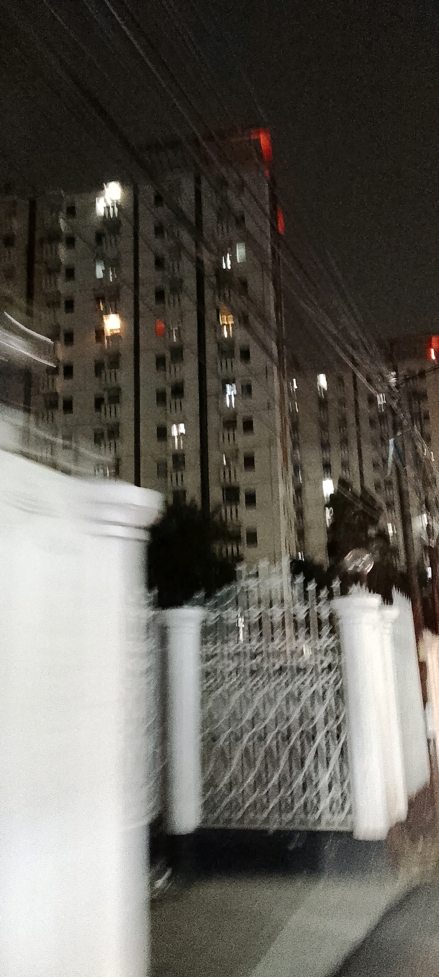 This is a night-time, motion-blurred photograph taken from a moving vehicle, likely in Depok, Indonesia, showing a high-rise apartment building. The building's facade is a grid of windows, with some windows illuminated, suggesting occupancy. A red light is visible near the top of the building, possibly a construction light or warning signal. Overhead power lines crisscross the sky. In the foreground, a white wall or fence with decorative pillars and a metal gate are also motion-blurred. The overall impression is one of urban nighttime activity with a slight sense of speed and movement.