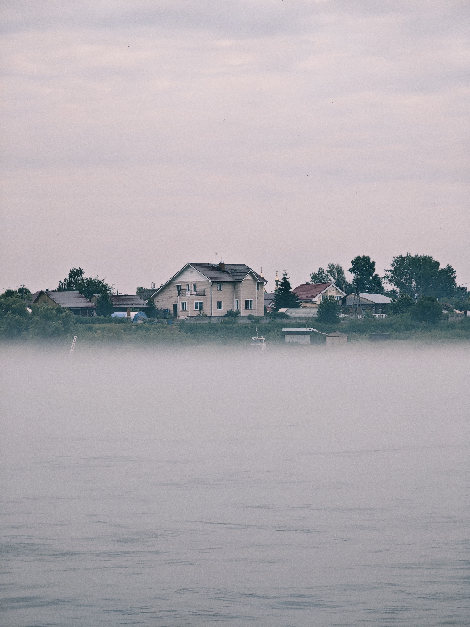 The image depicts a foggy morning scene on a body of water, likely the Angara River, near the city of Zheleznogorsk, Russia. A dense fog bank hovers low over the water, obscuring much of the foreground. In the background, a cluster of houses and trees are visible on the far bank, appearing hazy due to the fog and distance. The sky is overcast with a soft, pale light. A few small birds are seen flying in the sky. The overall atmosphere is serene and slightly melancholic, characteristic of a misty dawn.