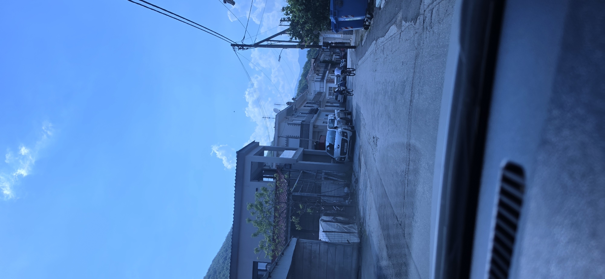 The image displays a street scene in Melivoia, Greece, captured from inside a vehicle, with the dashboard visible in the lower foreground. The time is daylight, under a clear blue sky with scattered cumulus clouds. The street is paved, exhibiting visible wear and patching. Two-story residential buildings with light-colored facades line both sides of the street, some featuring balconies and roof-mounted satellite dishes. A white Peugeot SUV, bearing Greek license plate YAN-5077, is parked on the left side of the street, followed by a lighter-colored pickup truck. In the middle distance, two individuals are riding bicycles away from the viewer down the street. On the right, a utility pole supports numerous overhead power lines, and two blue municipal waste bins are positioned on the sidewalk. Vegetated mountains form the background, indicating the town's geographic setting.