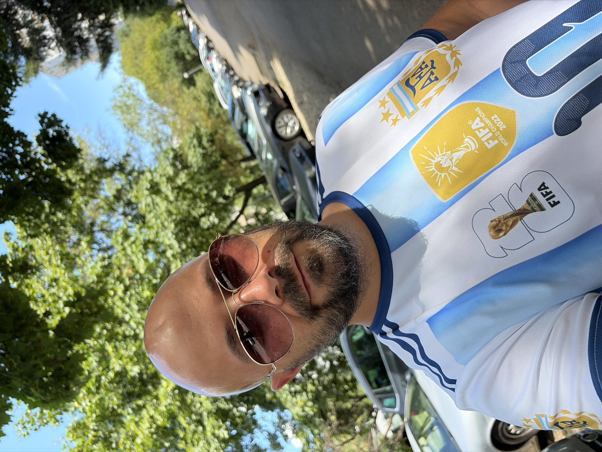 A bald, middle-aged man with a beard and sunglasses is taking a selfie outdoors. He is wearing an Argentina national football team jersey, featuring the FIFA World Cup champions 2022 badge and a trophy graphic. He is standing on a street lined with parked cars and trees. The man's head and torso are in the foreground, and the background shows a street with cars, trees, and a building. The scene is set in a city, possibly in Sofia, Bulgaria, indicated by the architecture and vegetation. The lighting suggests it is daytime.
