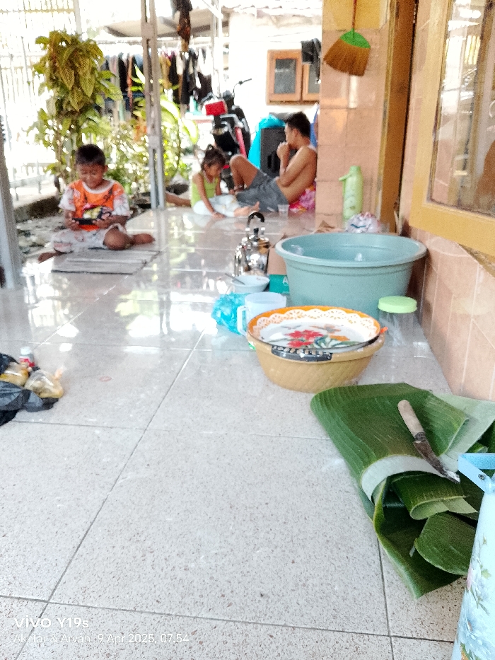 The image shows an outdoor patio in Gegesik, Indonesia. Three people are visible. One is an adult male sitting on the floor with his legs crossed, looking towards the right, and another is a young boy sitting on the floor with his legs crossed, looking down at a small, rectangular object. There is also a young girl wearing a pink shirt sitting on the floor, looking to the left. The adult male is wearing a dark gray t-shirt and the boy is wearing an orange and white shirt with a blue stripe. The girl is wearing a pink shirt and black pants. The patio has a light gray tiled floor, and there are numerous objects scattered around, including a small metal teapot, a blue plastic basin, a large wicker plate, and a stack of banana leaves. The banana leaves are next to a blue and white bucket. There are several other objects visible, including a white plastic bag, a white water bottle, and a knife on the banana leaves. The patio is situated next to a building that has a brown wall. The building has several large windows. A large green leafy plant is visible to the left of the patio, which may be part of a garden. The image was taken at 07:54 on April 9, 2025. There are several other objects in the image, including a white water bottle, a white plastic bag, and a knife.