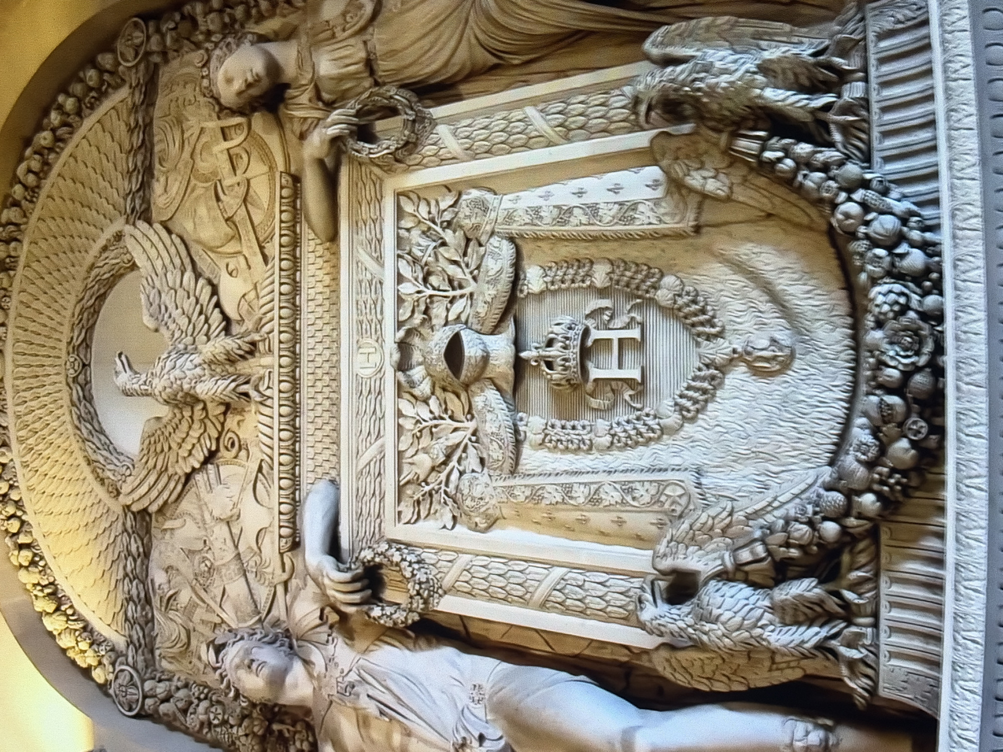 An ornate, carved stone relief depicts a central coat of arms framed by laurel branches and surmounted by a helmet and crown. Two large eagles flank the coat of arms. The coat of arms features a prominent stylized letter "H" below the crown. Above the coat of arms, a large eagle with outstretched wings dominates the composition. On either side of the central relief are life-sized, carved figures of women. The woman on the left reclines, draped in classical attire, and holds a wreath. The woman on the right stands, also in classical dress, and holds a wreath. The entire relief is framed by decorative elements, including a semi-circular element above the eagle and a base with further carved details. The relief appears to be part of a monument or architectural feature, possibly in an interior setting given the lighting and detail.