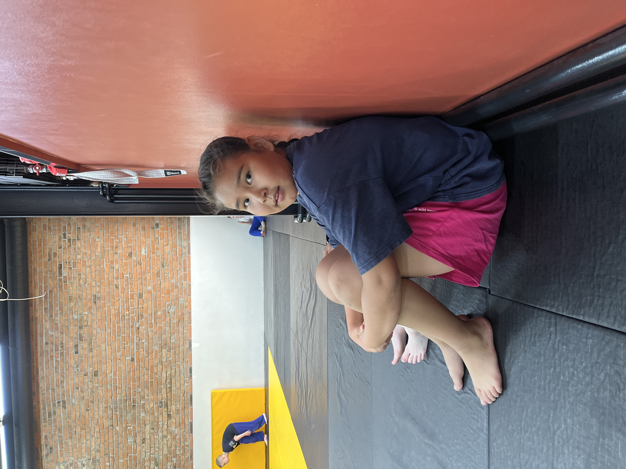 In an indoor gym with a brick wall and orange padded wall, a young girl with braided hair and a navy blue t-shirt and pink shorts sits on a grey mat, leaning against the orange wall. Her legs are crossed, and her arms are wrapped around her knees. Another person's bare feet are visible to her left. In the background, a boy in a blue uniform is practicing on a yellow mat, and another person in blue is sitting on the mat. The environment appears to be a martial arts or gymnastics facility. This scene is not specific to the city of Krasnoyarsk, Russia, as no identifying landmarks or context are present.