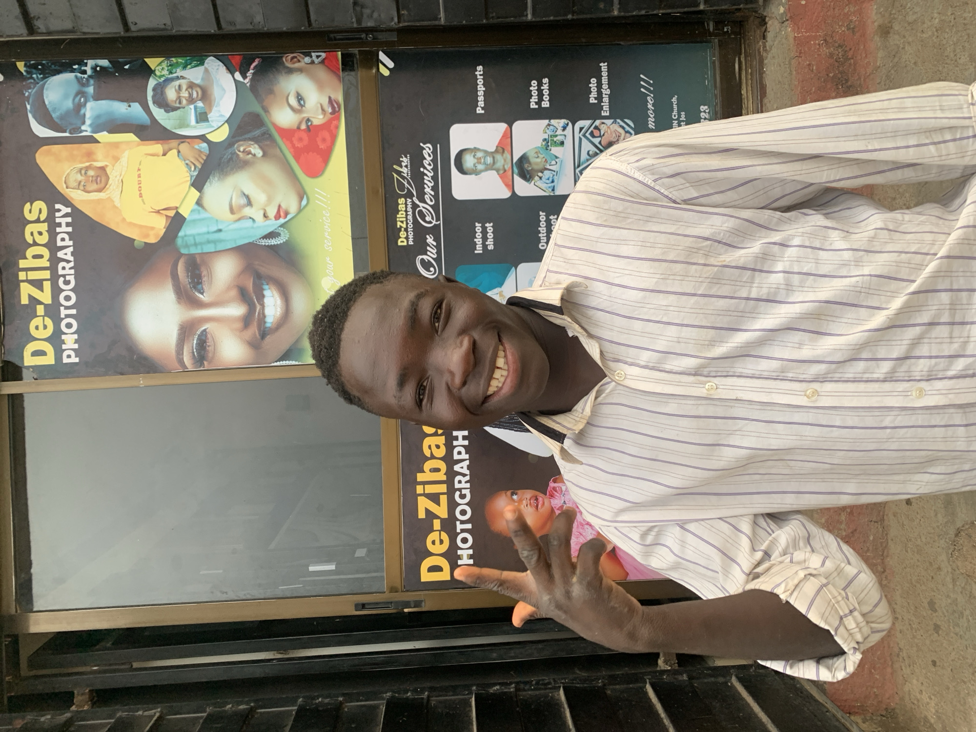 A man stands smiling in front of a photography studio called "De-Zibas Photography." The studio is located in Jos, Nigeria, specifically near "IN Church, et Jos," according to text on the signage. The man wears a light-colored striped shirt. Behind him, the studio windows display advertisements for various photography services, including passports, photo books, photo enlargements, indoor shoots, and outdoor shoots. Sample photos of people are also featured in the advertisements.