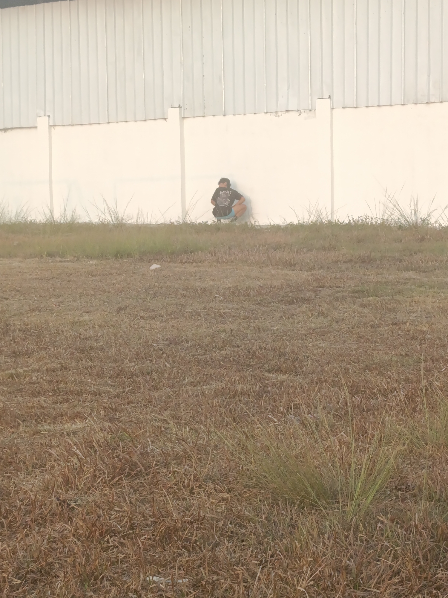In a dusty, overgrown field in Sumberejo, Indonesia, a person wearing a dark t-shirt and light-colored shorts is squatting near a beige wall. The wall is likely part of a building, as it is constructed of corrugated metal at the top and solid panels below. The person appears to be alone, with no visible interactions. The ground is covered in dry, brown grass, with some patches of green weeds. A piece of white litter is visible in the grass in the foreground. The overall impression is one of an outdoor, undeveloped area.