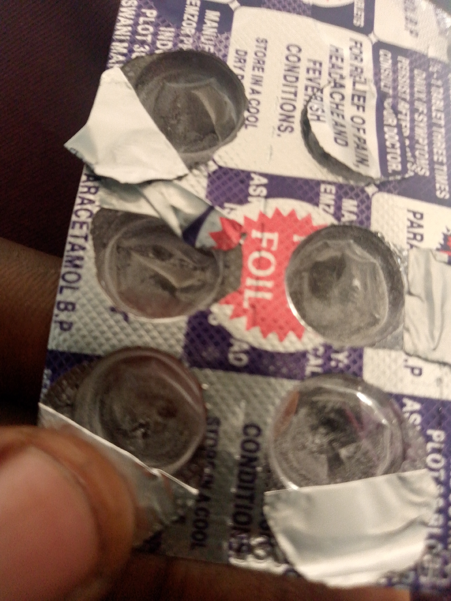 The image shows a blister pack of paracetamol tablets. The blister pack is partially opened, with several of the individual tablet cavities empty and the foil backing torn. Text on the blister pack includes "PARACETAMOL B.P.", "FOR RELIEF OF PAIN, HEADACHE AND FEVERISH CONDITIONS.", and "STORE IN A COOL DRY PLACE.". There are also some illegible markings, possibly manufacturing or lot numbers. A thumb and part of a finger are visible on the left side of the image, obscuring some of the blister pack. The background is dark and out of focus. There is no information or visual evidence in the image that would allow for identification of the location as Jauro Bamvo, Nigeria.