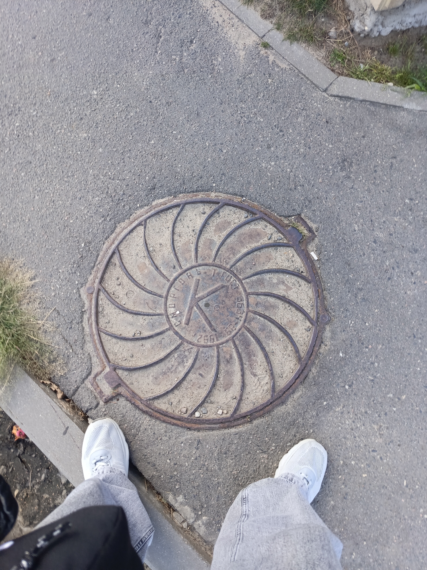 The image depicts a top-down view of a manhole cover in Ivatsevichy, Belarus. The manhole cover is circular and made of metal, with a decorative pattern resembling swirling lines radiating from a central "K" emblem. The year "1992" is also visible on the cover. The manhole cover is set into asphalt pavement, with a concrete curb and sparse grass to the left. The lower part of the image shows the feet of a person wearing light blue sneakers and gray jeans, suggesting they are standing near the manhole cover. A black bag is partially visible in the lower left corner.