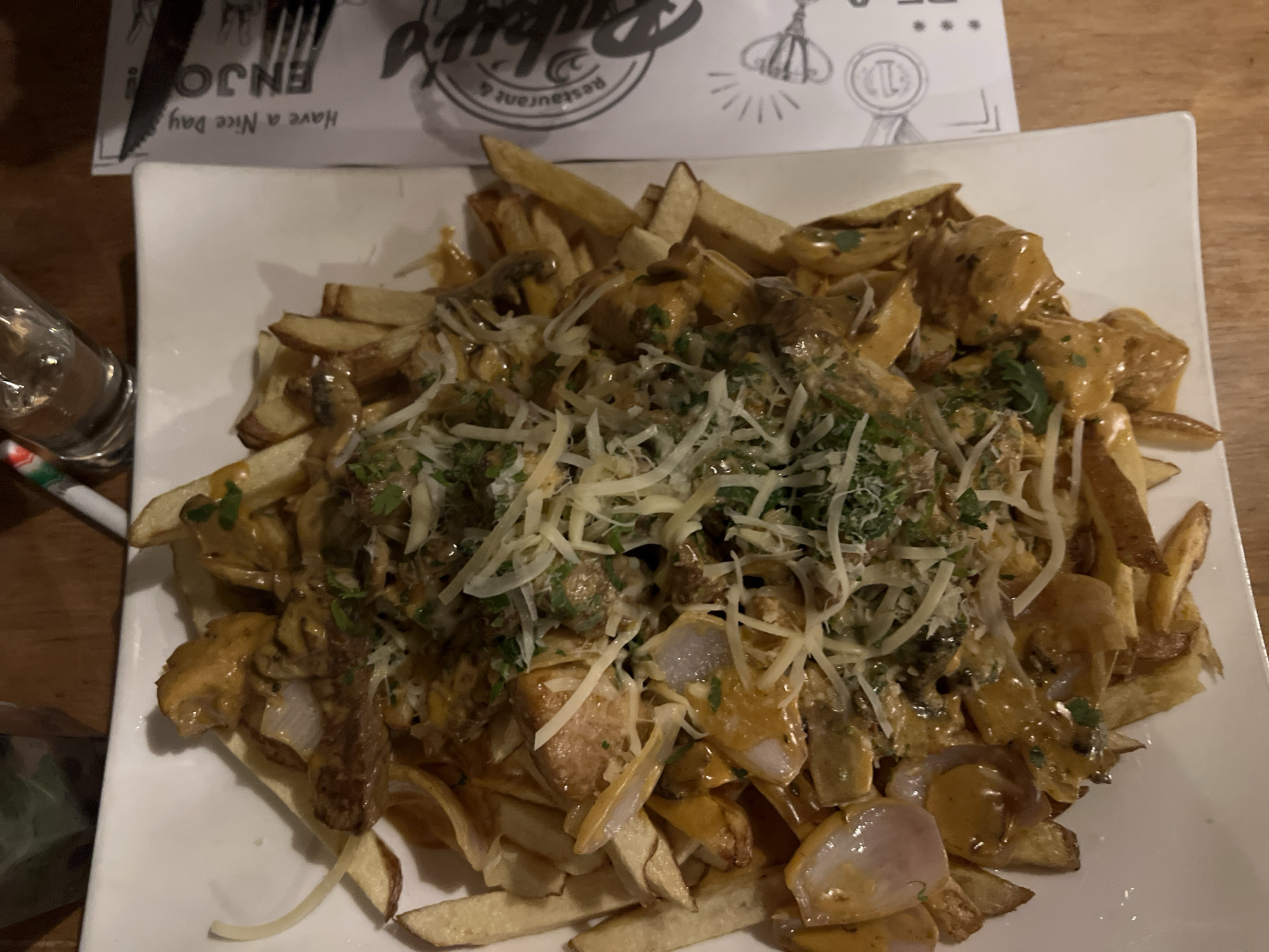 The scene depicts a close-up, overhead view of a prepared dish served on a white rectangular plate, situated on a dark wooden table in a restaurant in Arica, Chile. The plate holds a large portion of golden-brown, skin-on french fries. These fries are generously covered with a creamy, light brown sauce. Within the sauce and interspersed among the fries are irregular chunks of cooked protein, possibly meat, along with slices of mushrooms and segmented pieces of red onion. The entire dish is garnished with a substantial amount of finely shredded white cheese and sprinkled with chopped green herbs. To the upper left of the plate, a clear drinking glass is partially visible on the table, accompanied by a white object featuring red and green stripes, potentially a straw or stirrer. Above the plate, a white sheet of paper, likely a menu or placemat, displays partial text including "ENJO !" and "Have a Nice Day," along with other graphical elements and the phrase "Restaurant &".