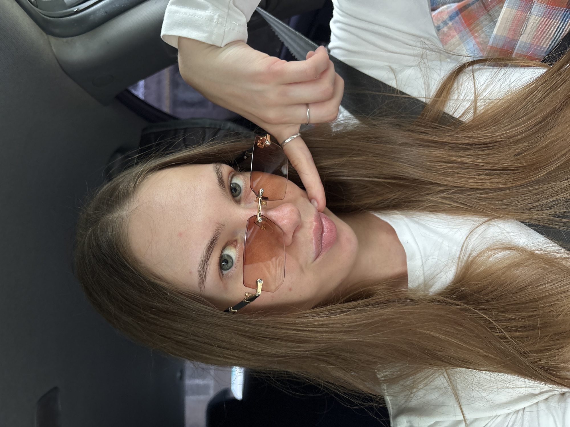 A young woman with long, light brown hair and fair skin is shown in a close-up, tilted shot. She is wearing a white long-sleeved shirt and a plaid shirt over it. She has on large, rimless, amber-tinted sunglasses with gold accents. Her right hand is raised, with one finger touching her cheek. She is wearing two delicate silver rings on her left hand. The background suggests she is inside a car, with a portion of the dark gray interior visible. The lighting appears to be natural, possibly daylight, and there are no strong shadows, suggesting it might be overcast or the sun is not directly visible. The woman's expression is a playful, slightly pensive look towards the camera. There is no visible text in the image. The location context provided is Perm, Russia.