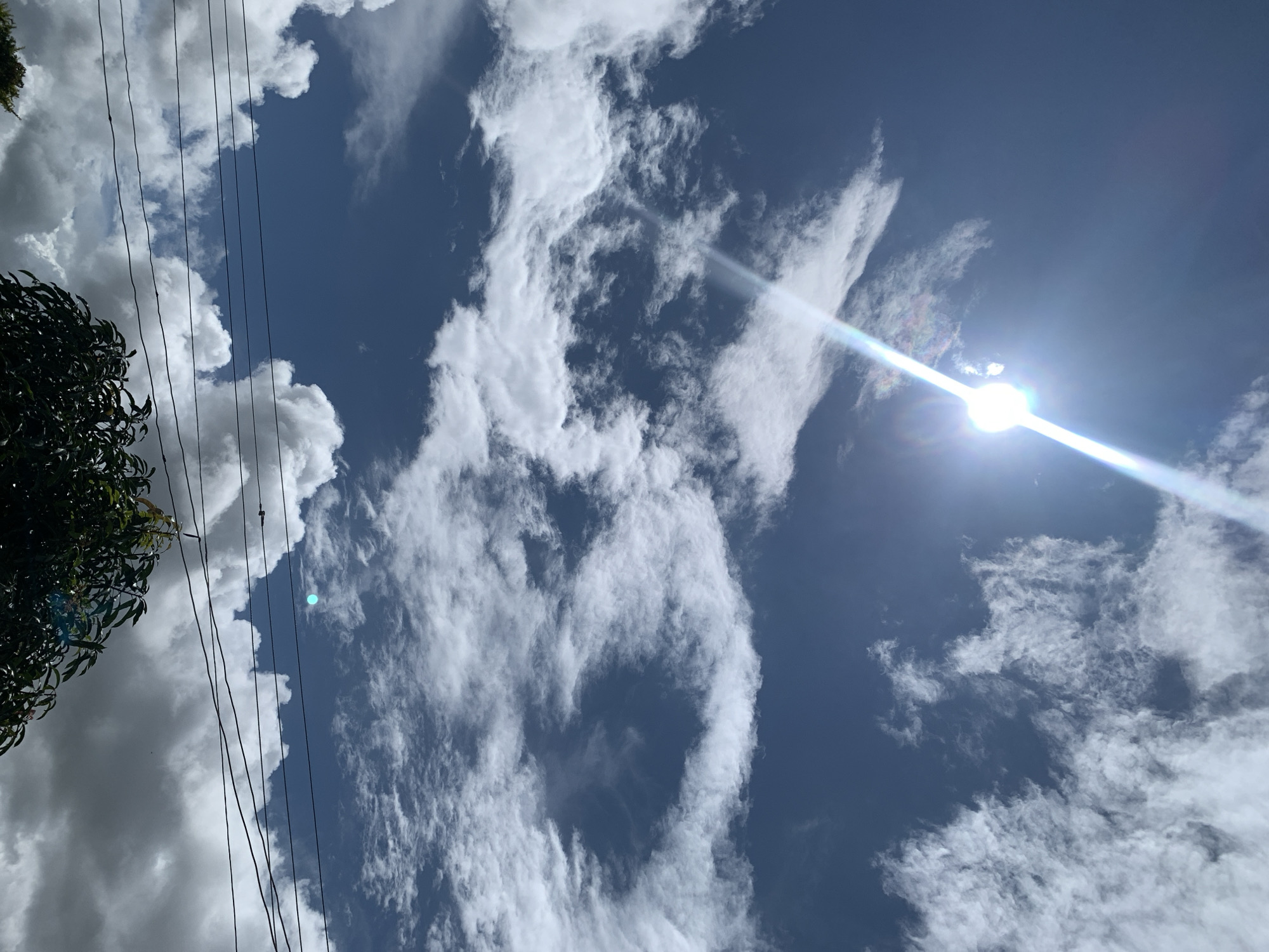 The image depicts a bright, sunny day with a predominantly blue sky filled with a variety of white clouds. The sun is prominently visible in the upper left quadrant, emitting a strong glare with a rainbow-like effect. Wispy, thin clouds are scattered across the sky, interspersed with larger, more voluminous cumulus clouds. In the lower portion of the image, a dense green tree with dark leaves is visible, suggesting an outdoor setting. Several black electrical wires run horizontally across the scene, adding a man-made element to the natural landscape. The overall impression is one of a clear day with dynamic cloud formations. The location of Kuru Karama, Nigeria, is not discernible from the visual elements of the image itself.