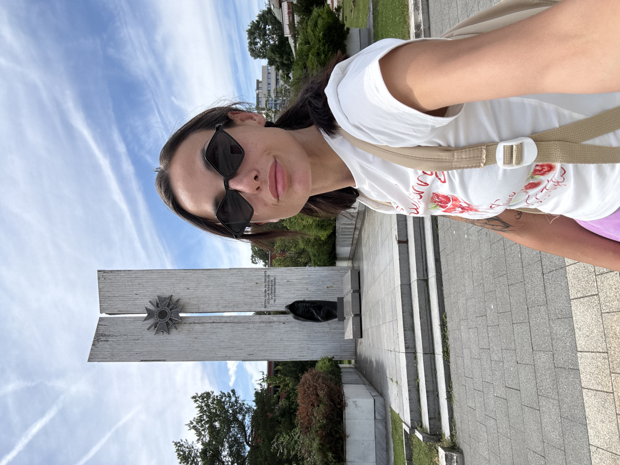 A young woman with dark hair, wearing sunglasses and a white t-shirt with a strawberry print, takes a selfie in front of the Pantheon of Revival Masters in Pleven, Bulgaria. The monument, a tall, imposing structure with two slanted concrete pillars, features a star-shaped emblem at the top and a dark, hooded sculpture at its base. The scene is set on a sunny day with a partly cloudy sky, surrounded by greenery and paved walkways. The Pantheon is a significant historical monument in Pleven, commemorating Bulgarian national heroes.