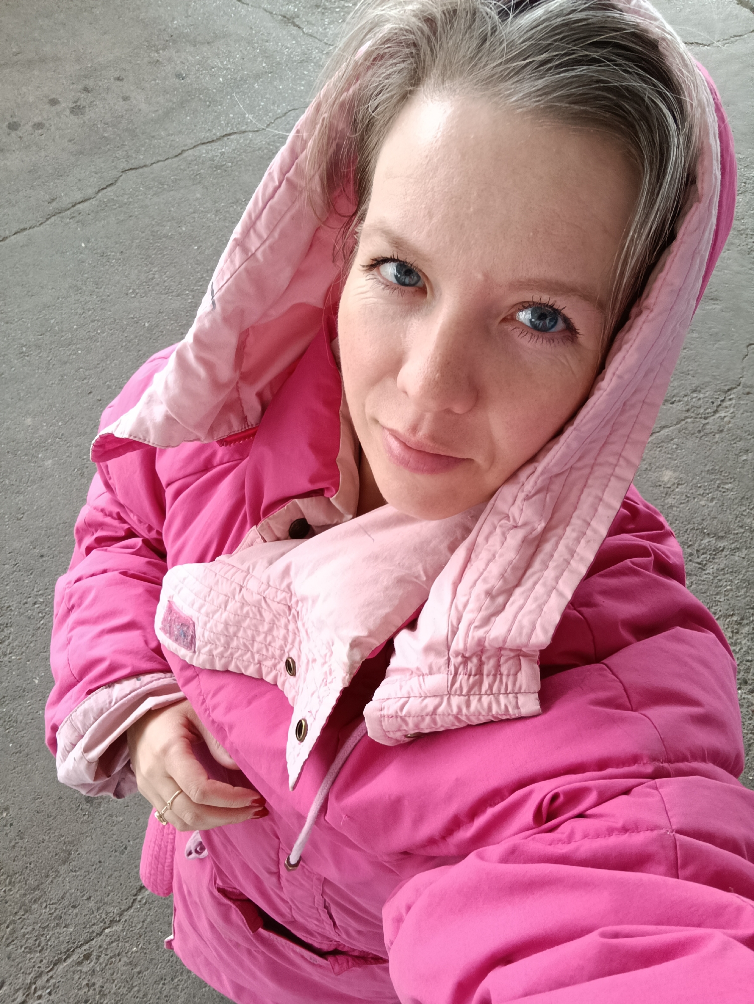 A close-up, high-angle portrait features a woman with blue eyes and light skin, looking directly at the camera with a neutral to slightly smiling expression. Her hair, which appears to be light-colored with some visible gray strands, is partially covered by a hood. She is wearing a bright, almost fuchsia pink, padded or quilted winter jacket. The jacket has a contrasting lighter pink lining or collar visible around her neck and inside the hood. Her left hand is visible near the bottom of the frame, showing red painted fingernails and a ring with a small charm on her ring finger. The background consists of gray, cracked pavement or concrete, suggesting an outdoor setting. No specific time of day is discernible beyond daylight, and the jacket suggests cooler weather. There is no visible text in the image. The photo was taken in Chaikovsky, Russia.
