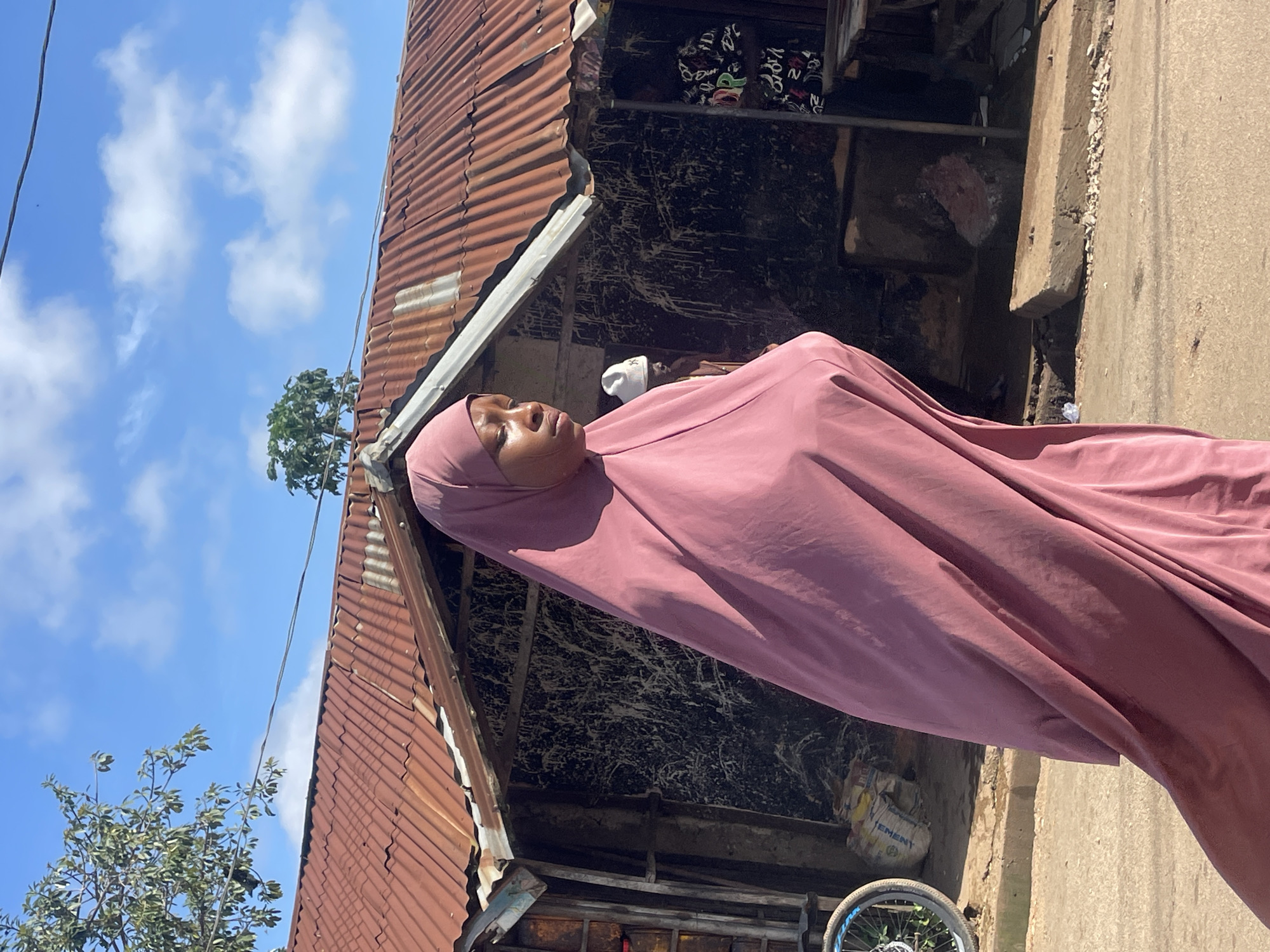 An outdoor, eye-level, medium shot captures a young woman in a rose-pink hijab walking from left to right, her gaze directed downward and to the right. She is positioned in the foreground, with her body angled away from the viewer and her hijab flowing around her. In the mid-ground, a weathered, corrugated metal roof of a market stall is visible, casting a shadow over the dark, textured wall behind it. A man wearing a white beanie is seen in the background to the left of the woman. To the far left, a bicycle wheel with blue accents is partially visible. The environment appears to be a street in Jos, Nigeria, characterized by a dusty, unpaved surface and a rustic, open-air market stall. The sky above is bright blue with scattered white clouds, indicating daytime.