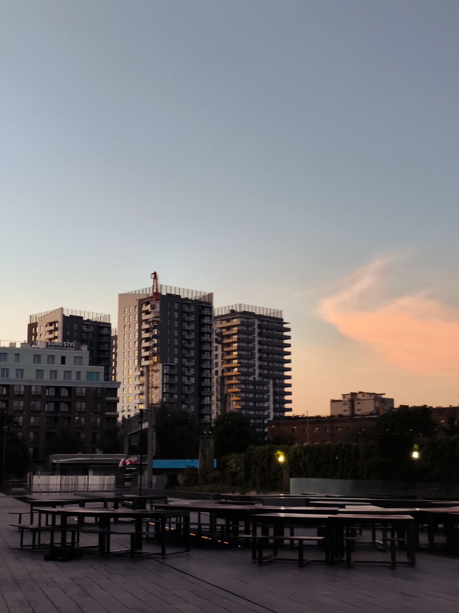 This is a medium shot, taken from a low angle, of an outdoor seating area in Gdańsk, Poland, in the early evening. The sky is a gradient of pale blue to a soft orange near the horizon, with a prominent, wispy cloud formation catching the sunset light.
In the foreground, there is a wooden deck with multiple dark, rectangular picnic tables with attached benches. The tables are arranged in rows, and some are empty, while others may have items on them, but they are too obscured by shadow to be identified.
In the midground, there are several modern, multi-story apartment buildings. The closest building on the left is lighter in color with visible windows. To its right, a taller, darker building stands, featuring a red construction crane at its peak. Further to the right, another similar dark apartment building is visible, its windows reflecting the warm evening light. Behind these buildings, there are hints of other structures and trees.
In the background, a line of darker, lower buildings stretches across the horizon, with a few lights visible. The overall impression is one of urban development and an evening atmosphere in a waterside or boardwalk area. There are no people clearly visible in the frame.