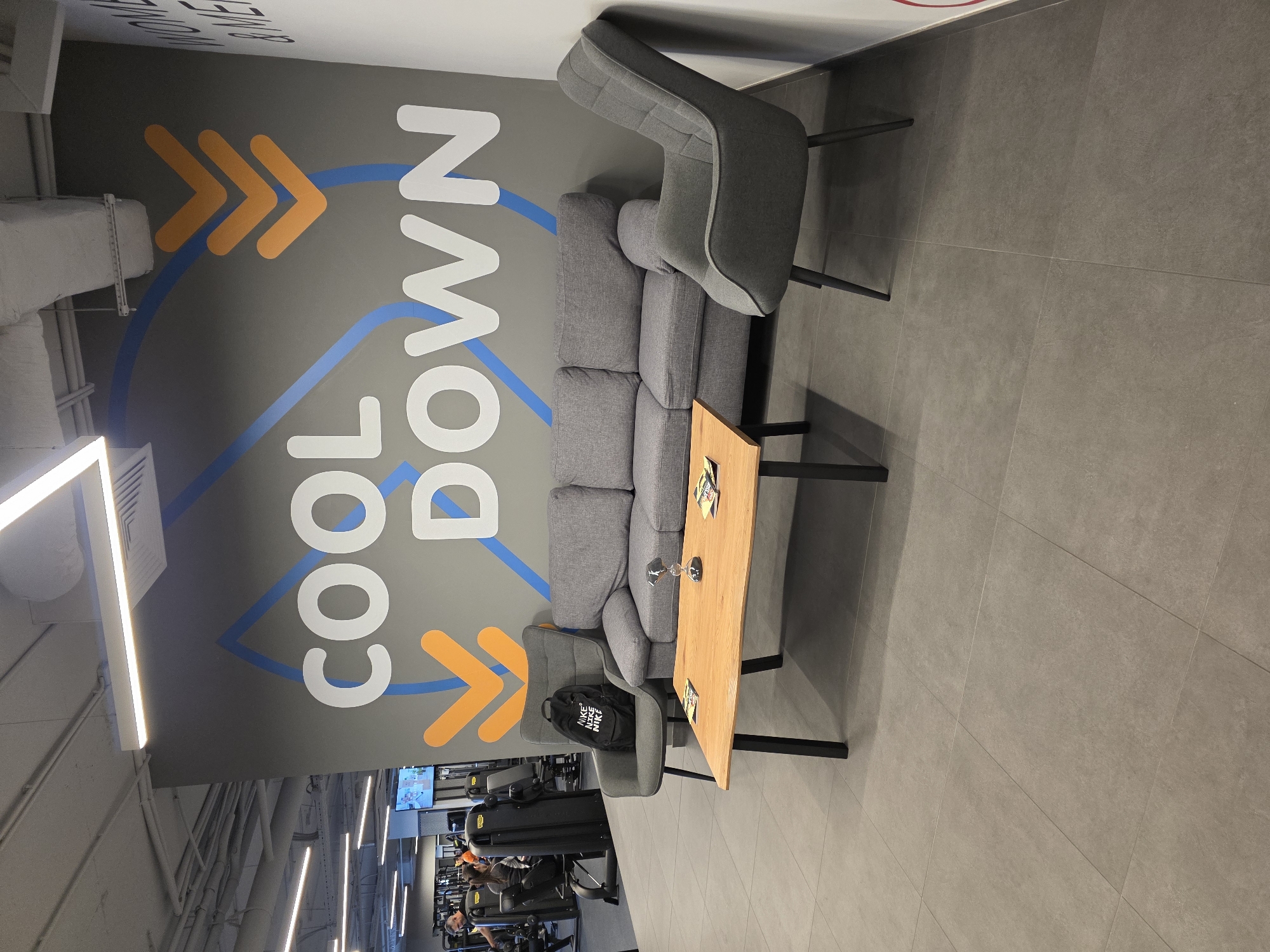 The scene is an interior shot of a modern gym. A gray wall features a "COOL DOWN" graphic in white letters with blue and orange arrow-shaped accents. In front of the wall, there's a gray sofa with a low, rectangular wooden coffee table. A gray chair sits at each end of the sofa. A black Nike backpack rests on one of the chairs. A glass and some papers are on the table. In the background, various gym equipment and people exercising are visible. The flooring is made of large gray tiles. Based on the presence of a gym facility and the general design aesthetic, the location is likely in a modern fitness center in Plovdiv, Bulgaria.
