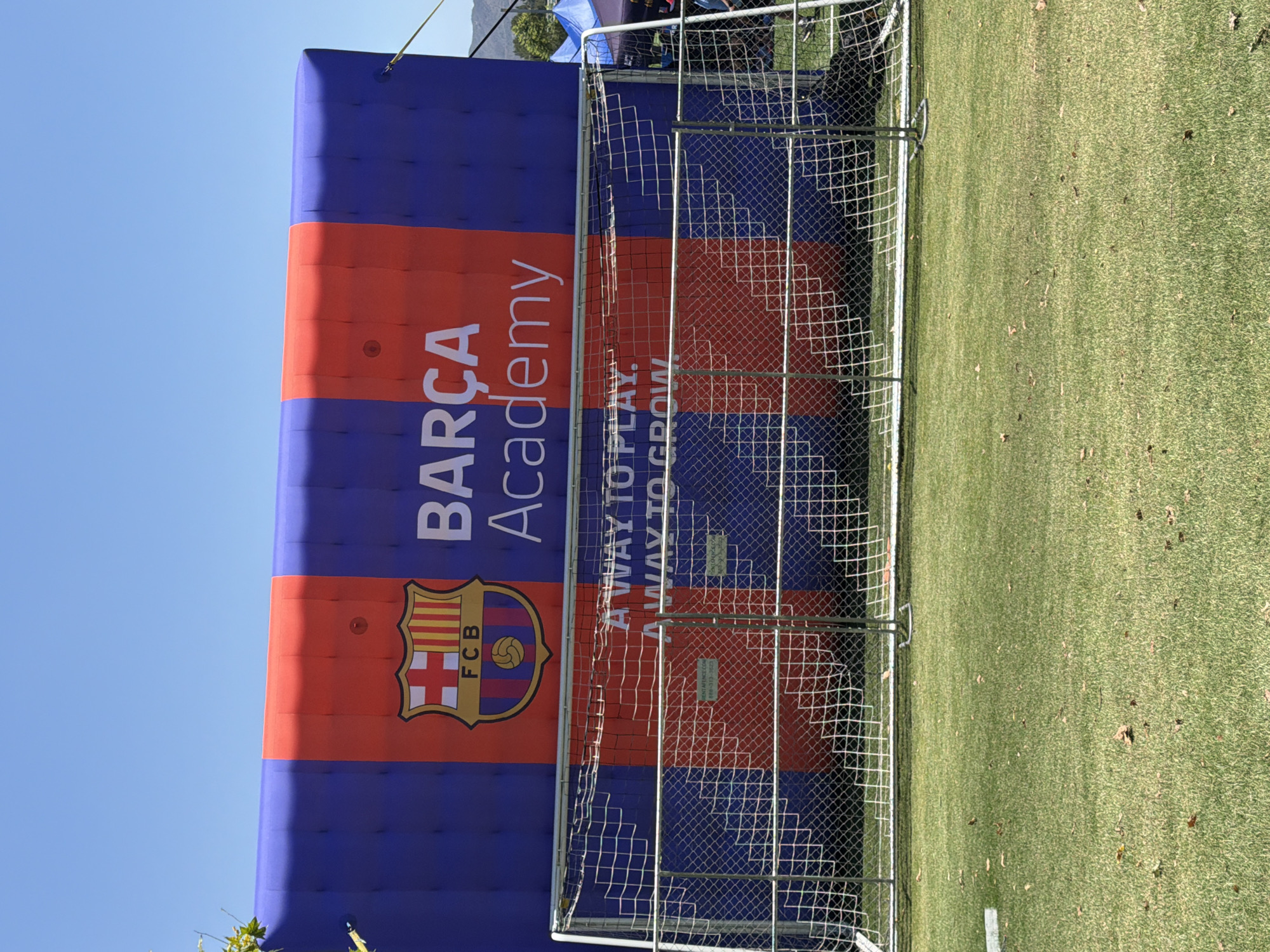 The image shows an inflatable structure with the "Barça Academy" logo and text, set up on a grassy field in front of a soccer goal. The structure is predominantly blue and red, with the Barça crest prominently displayed. The text "A WAY TO PLAY. A WAY TO GROW." is visible on the structure below the logo. A metal fence encloses the soccer goal. The sky is clear and blue, suggesting a daytime outdoor event. There are no people clearly visible in the frame. The location is identified as Irvine, California, United States of America.