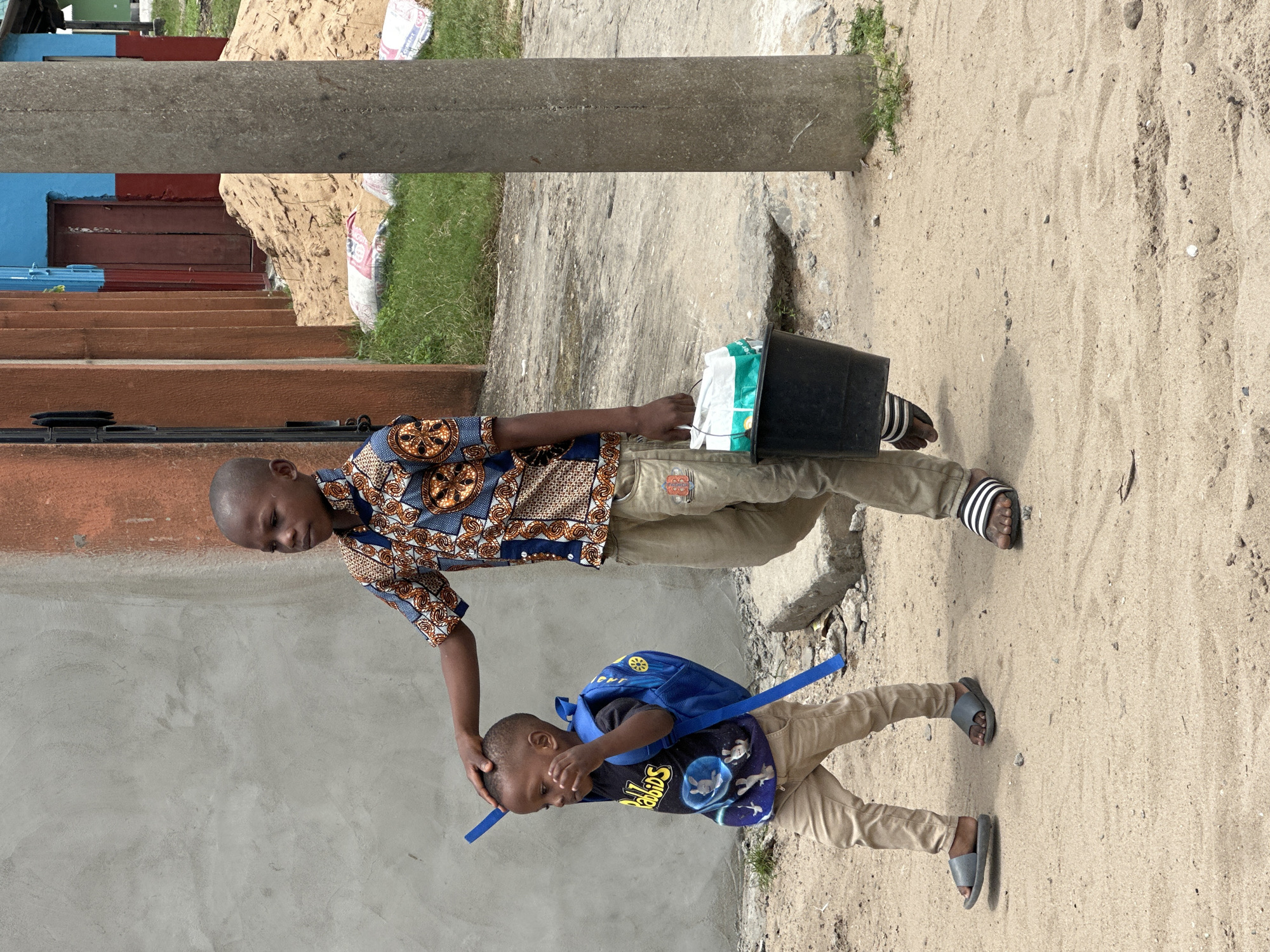 In Badagry, Nigeria, two young boys walk along a sandy path next to a building with a concrete and orange facade. The taller boy, on the right, wears a patterned shirt and light-colored trousers. He carries a black bucket with white and green items inside. The younger boy, on the left, has a blue backpack and wears a dark t-shirt with a graphic and light-colored trousers. He holds a blue object and walks with his head down. A concrete pillar stands to the right of the boys. In the background, a pile of sand and bags are visible next to a blue and red building.
