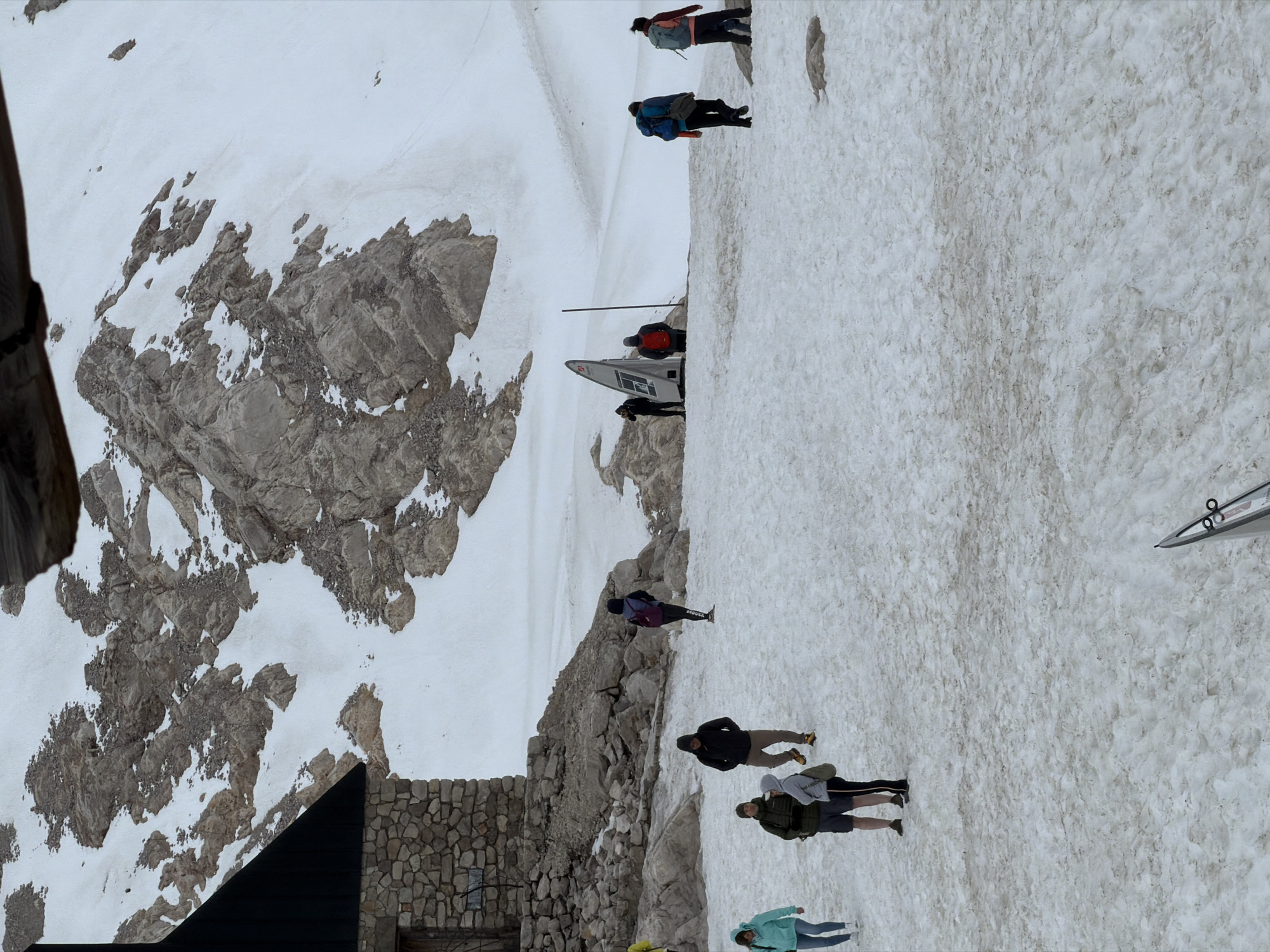 This is a medium-range shot of people in a snowy mountain environment near a stone and dark-roofed building. The building appears to be a mountain hut, possibly at the Zugspitze, Germany's highest peak, accessible from Garmisch-Partenkirchen. The ground is covered in patchy snow, with visible rocks and some brown dirt mixed in. Several individuals are scattered across the scene, most of whom appear to be hikers or tourists.
In the foreground, a person wearing a light blue jacket and jeans is walking towards the left. To their right, a group of three people are standing together. One is wearing a dark green jacket and shorts, another a light grey hooded top, and the third is wearing a light brown hooded jacket. Further back, a person in a dark hooded jacket is walking towards the right.
In the middle ground, on the right side of the frame, a person in a dark jacket with a purple backpack is walking away from the viewer. Further in the distance, near a modern-looking information kiosk, are two individuals, one wearing a red jacket and another in a dark outfit. Two more people are walking away from the kiosk towards the right, one with a blue backpack and another with a light blue backpack.
The background is dominated by a steep, rocky mountain slope partially covered in snow. The sky is overcast and a wooden overhang is visible at the very top of the image. The overall impression is a cold, possibly windy, mountain landscape with a few visitors.