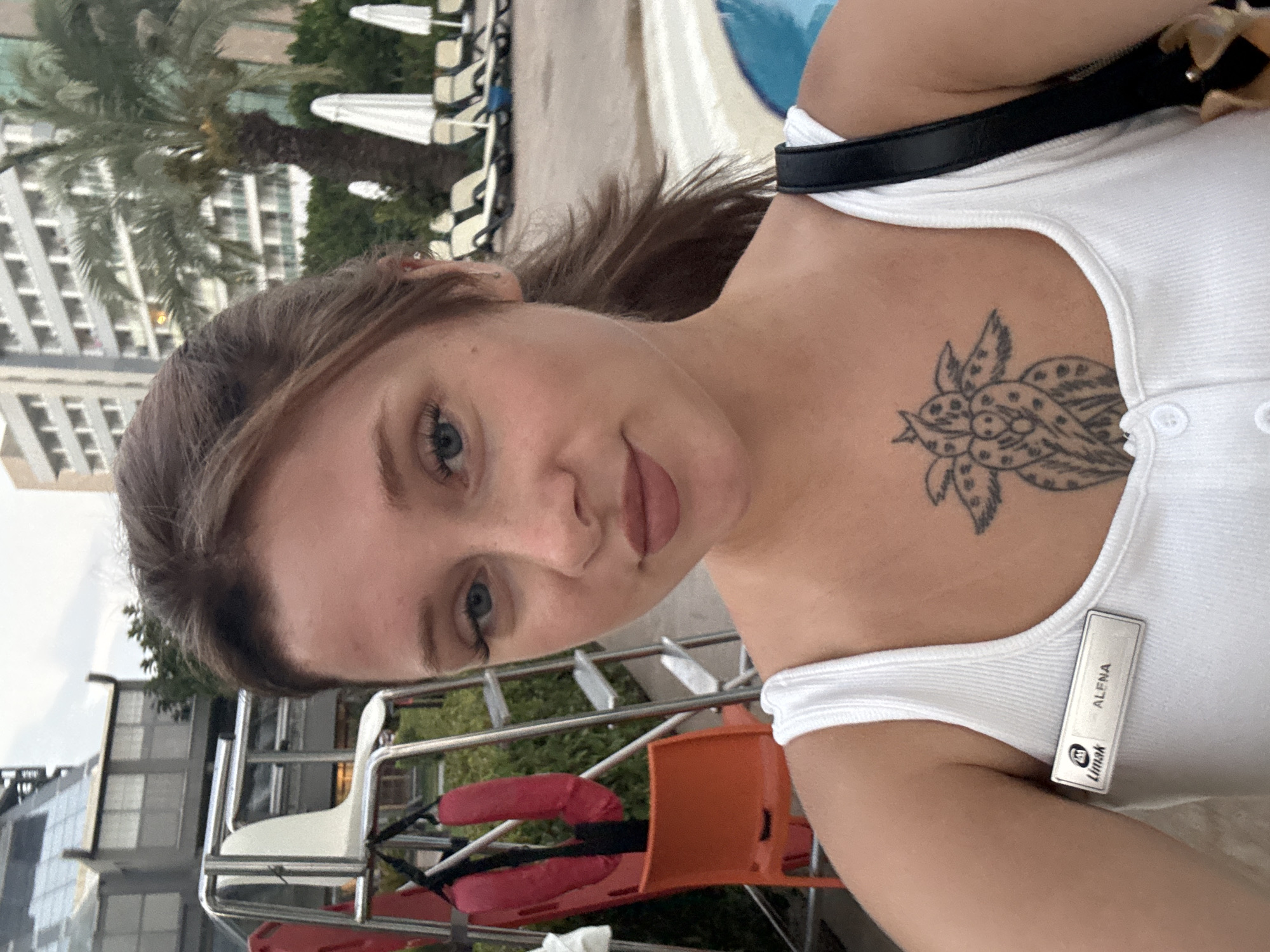 A young woman with light skin, blue eyes, and brown hair is taking a selfie outdoors. She is wearing a white tank top with a name tag that reads "Limak ALENA". She has a tattoo of a floral design on her chest. In the background, there is a swimming pool, lounge chairs, umbrellas, palm trees, and a multi-story building. There is also a lifeguard chair with red flotation devices and an orange chair. The location is Aksu, Turkey.