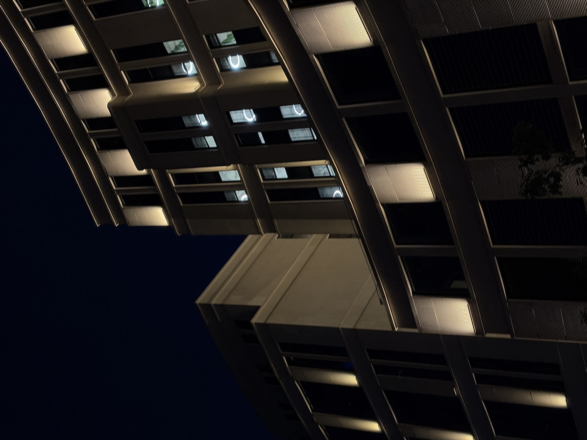 The image shows two modern buildings at night. The building in the foreground has a curved facade with many windows. The windows are dark, but some have reflections of circular lights. The building is illuminated by warm, yellow lights along its edges and on vertical sections. To the left, a second, lighter-colored building is visible. It also has windows and is lit by vertical strips of light. The sky is a deep, dark blue, indicating it is nighttime. The architectural style suggests a contemporary urban setting, possibly an office building or residential complex. The scene is devoid of people or any discernible activity. Given the context, these buildings are likely located in Minsk, Belarus.
