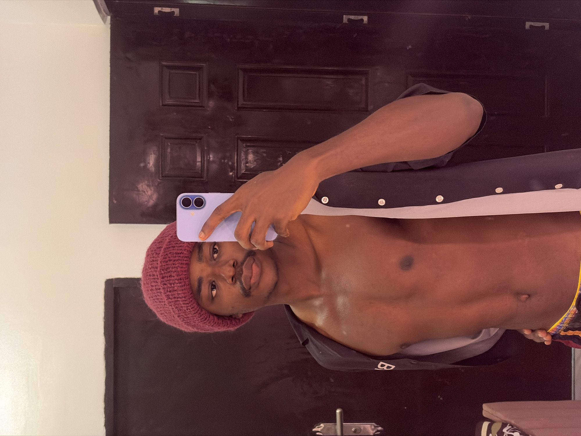 A man wearing a burgundy knit cap and an open dark blue shirt with white trim and buttons is taking a selfie with a purple smartphone. He has a mustache and his torso is bare. The background consists of a dark brown door with a rectangular panel design and a white wall. The location is Igboefon, Nigeria.