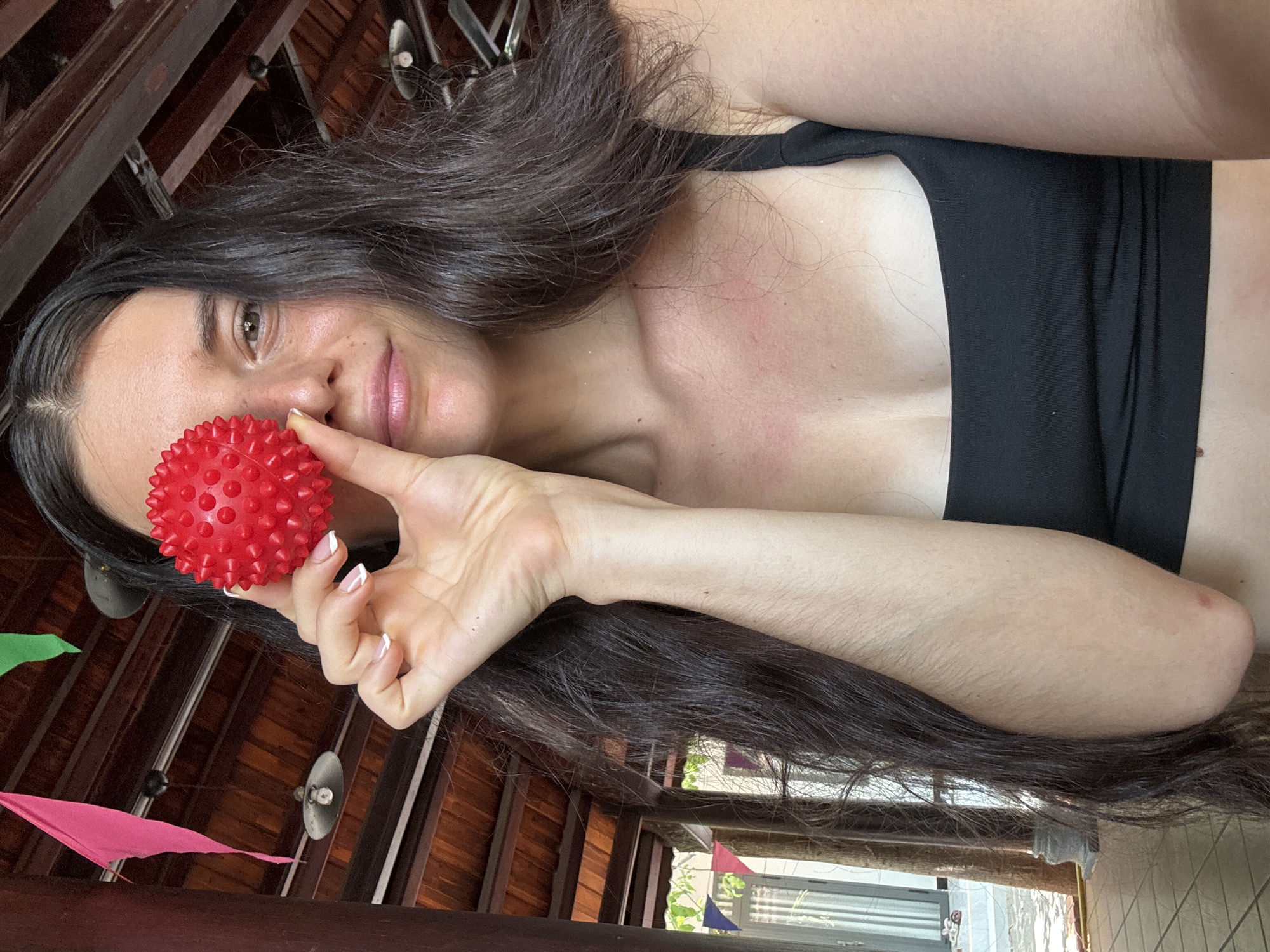 A young woman is pictured from the chest up, holding a small, red, spiked ball in front of her right eye. She has long, dark hair that falls over her shoulders and chest, and she is wearing a black sports bra. The background appears to be an outdoor covered area with a wooden ceiling and exposed beams. Red and green triangular flags hang from the ceiling on the left side of the image. Sunlight filters in from the left, creating shadows and highlighting the textures of the wooden structure. The scene suggests a casual, possibly resort-like setting. The location is Mũi Né, Vietnam.
