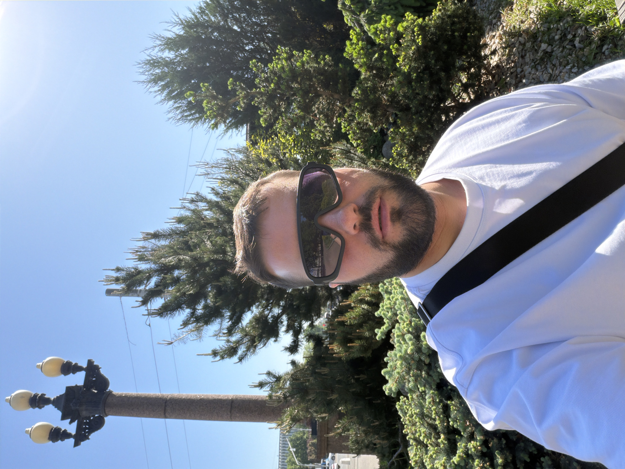 A man with a beard and sunglasses is taking a selfie outdoors on a sunny day. He is wearing a white shirt and a black strap, possibly from a bag, is visible across his shoulder. Behind him are trees and a light blue sky. To the left, there is a lamp post with four lights. Based on the architectural style of the lamp post and the presence of coniferous trees, the location is likely Moscow, Russia.