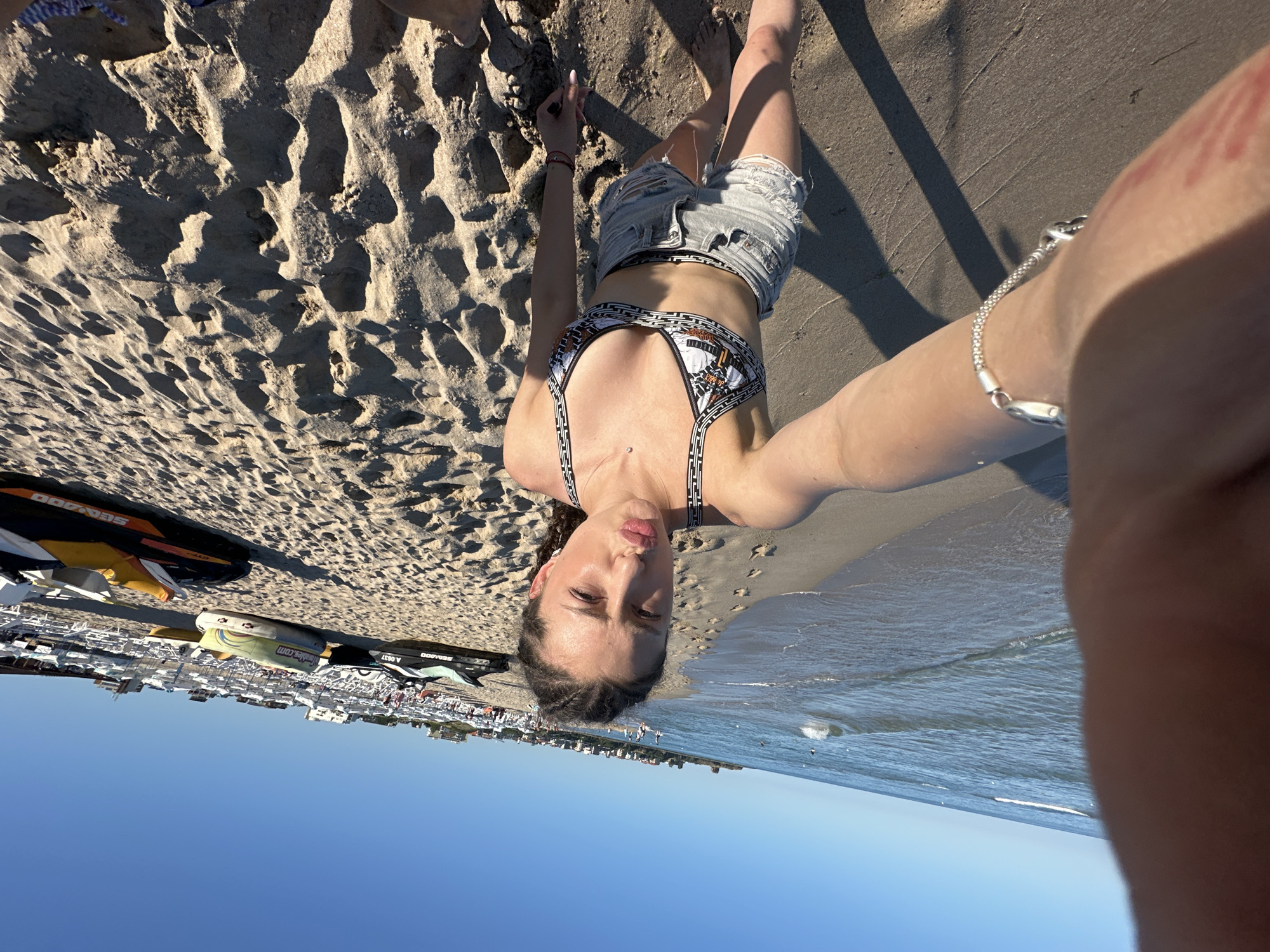 A young woman with braided hair is taking a selfie on a sandy beach in Primorsko, Bulgaria. She is wearing a patterned bikini top and distressed denim shorts. The sea is visible in the background with gentle waves. Numerous beach chairs and umbrellas are set up on the sand, along with several jet skis parked further up the beach. The sky is clear and blue.