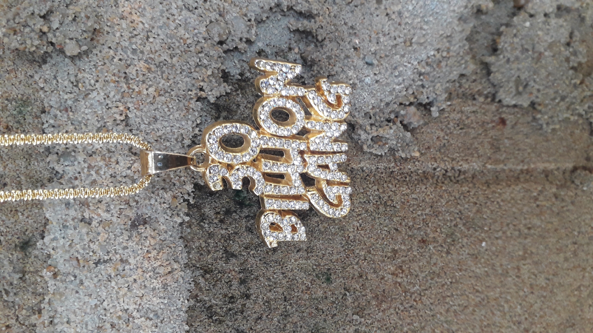 A gold-colored chain necklace with a pendant that reads "Big Don Ofo Nu" is lying on a rough, sandy surface. The pendant and chain are adorned with clear, shiny stones, possibly diamonds or crystals. The background appears to be a weathered, grainy surface, possibly concrete or compacted soil. No people are visible. The location in Nkpor, Nigeria is assumed, but not evident in the image itself.