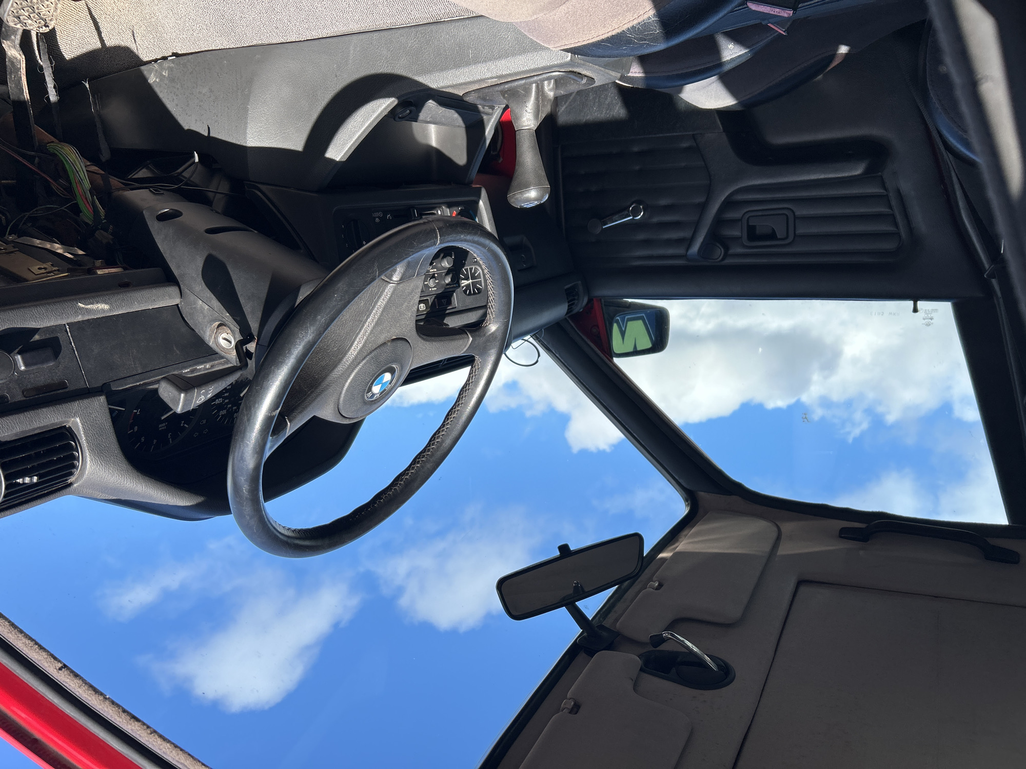 The image shows the interior of a car, specifically the driver's side dashboard, steering wheel, and front window, with a view of a bright blue sky with white clouds visible through the windshield. The car appears to be in a state of partial disassembly, with exposed wiring under the dashboard. The rearview mirror is visible. The car's interior is a mix of black and light brown/beige. The location in the city of Rutherglen, United Kingdom cannot be determined from the given image.
