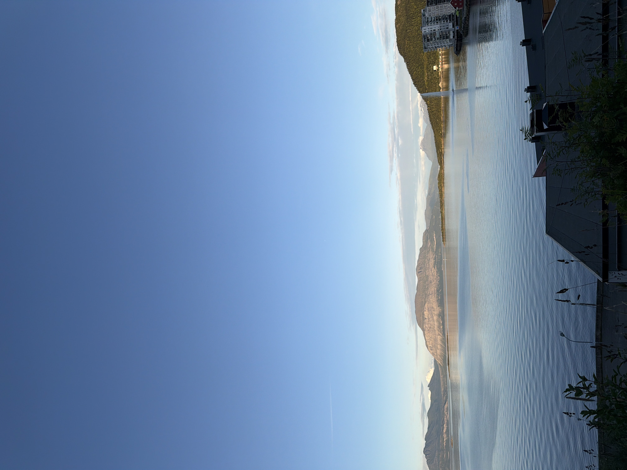 This image captures a serene waterfront view in Harstad, Norway. The foreground features the dark rooftops of buildings, with glimpses of lush green vegetation and what appear to be streetlights. The midground is dominated by a vast, calm body of water reflecting the clear blue sky and the surrounding landscape. A prominent fountain shoots a tall stream of water into the air, creating a focal point on the water's surface. In the distance, majestic mountains rise, some with snow-capped peaks, under a bright, expansive sky with a few wispy clouds. The scene suggests a peaceful day in a picturesque Norwegian city.