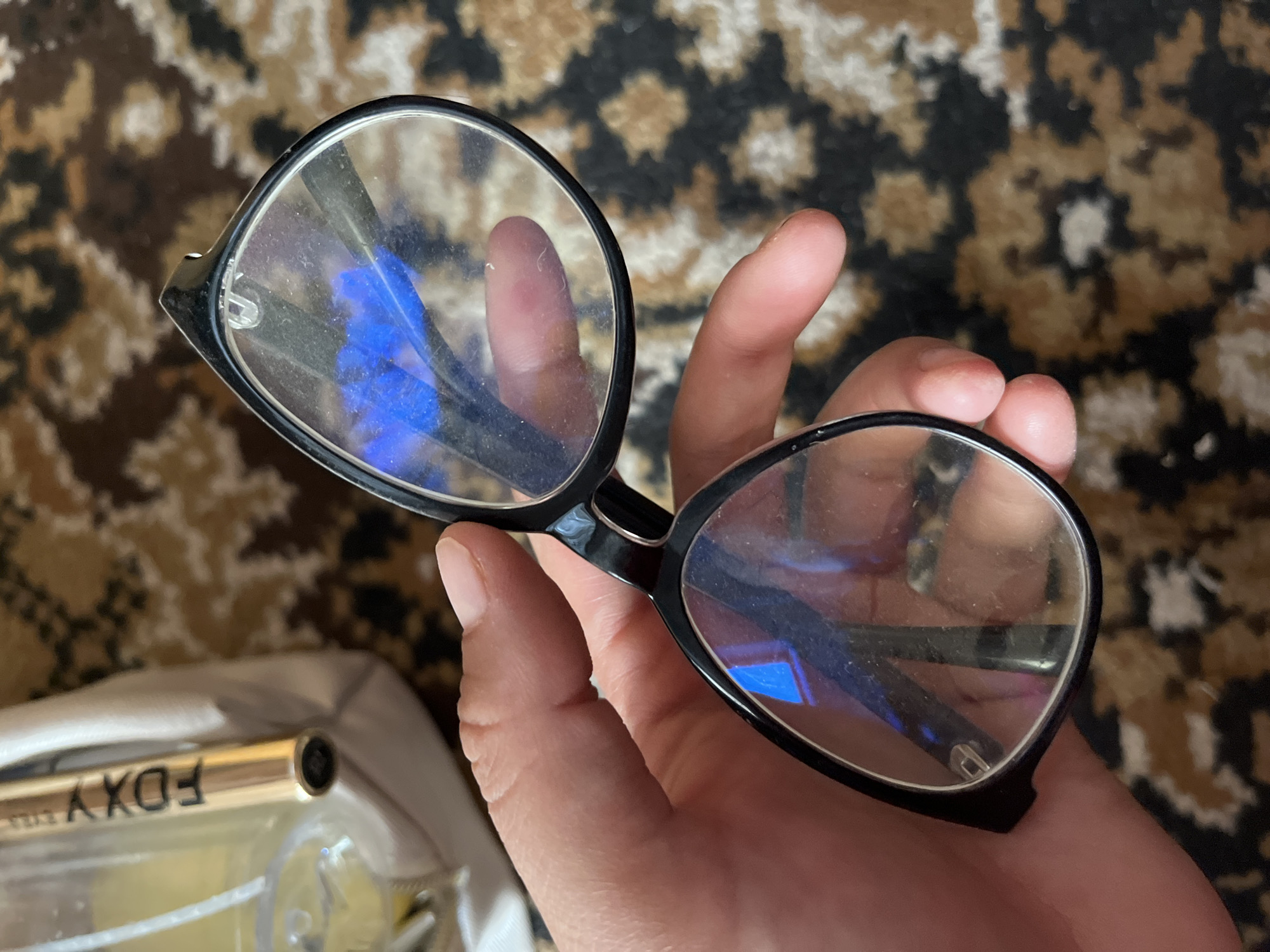 A person's hand holds a pair of black-rimmed eyeglasses, the lenses reflecting a blue light. The eyeglasses are black and have a slightly cat-eye shape. The hand is positioned to hold the glasses by the right arm and the top of the frame. The background is a patterned carpet in shades of brown and beige. In the upper left corner, part of a light-colored bag and a gold-colored tube with the word "FOXY" printed on it are visible. The overall scene appears to be an indoor setting.