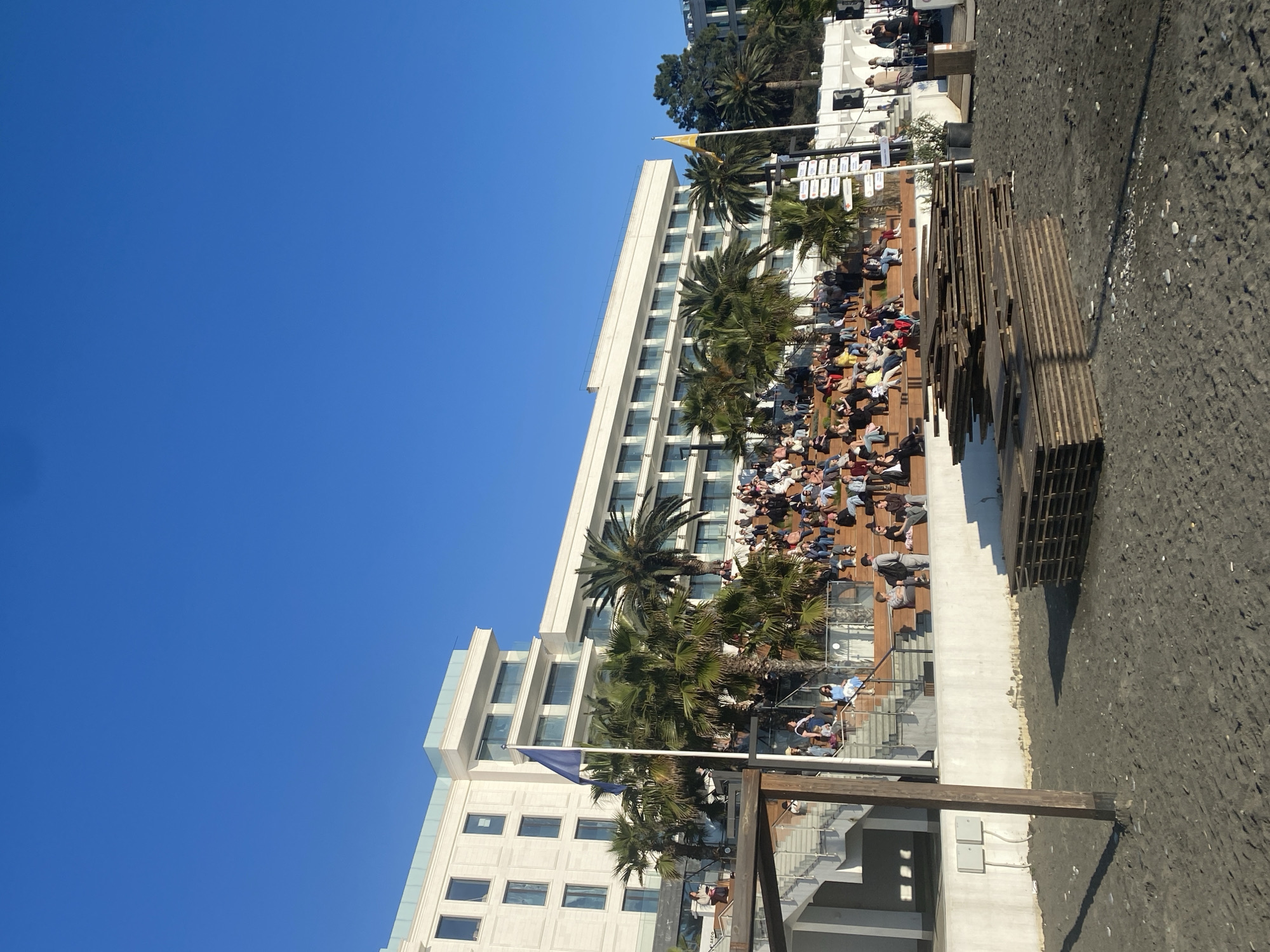 The image depicts a coastal scene in Sochi, Russia, featuring a large, modern white building, likely a hotel, with multiple stories and numerous windows. Palm trees are interspersed along the building's façade. An outdoor tiered seating area is visible, populated with people sitting in chairs, apparently watching or attending an event. The area abuts a dark, sandy beach. In the foreground, there are some stacked wooden structures, perhaps related to the beach or seating area. Above, there is a clear blue sky.