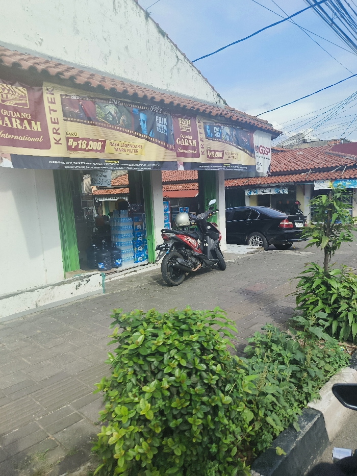 The scene shows the front of a small store in Purwakarta, Indonesia, located at coordinates -6.545 latitude and 107.442 longitude. The store has a white facade, a brown tiled roof, and a large sign advertising "Gudang Garam International" and "Kreteks" with a price of Rp. 18,000. The sign is attached to the front of the store, which appears to sell beverages and other products. A red motorcycle is parked in front of the store. A black car is parked on the side of the street in front of the store. The store's windows are covered with green, slatted blinds. There are several green plants in the foreground of the scene. A small section of the street is visible in the foreground. The sky is clear blue, with some wispy clouds. The street and sidewalk are paved with grey concrete blocks. Power lines run across the top of the image.