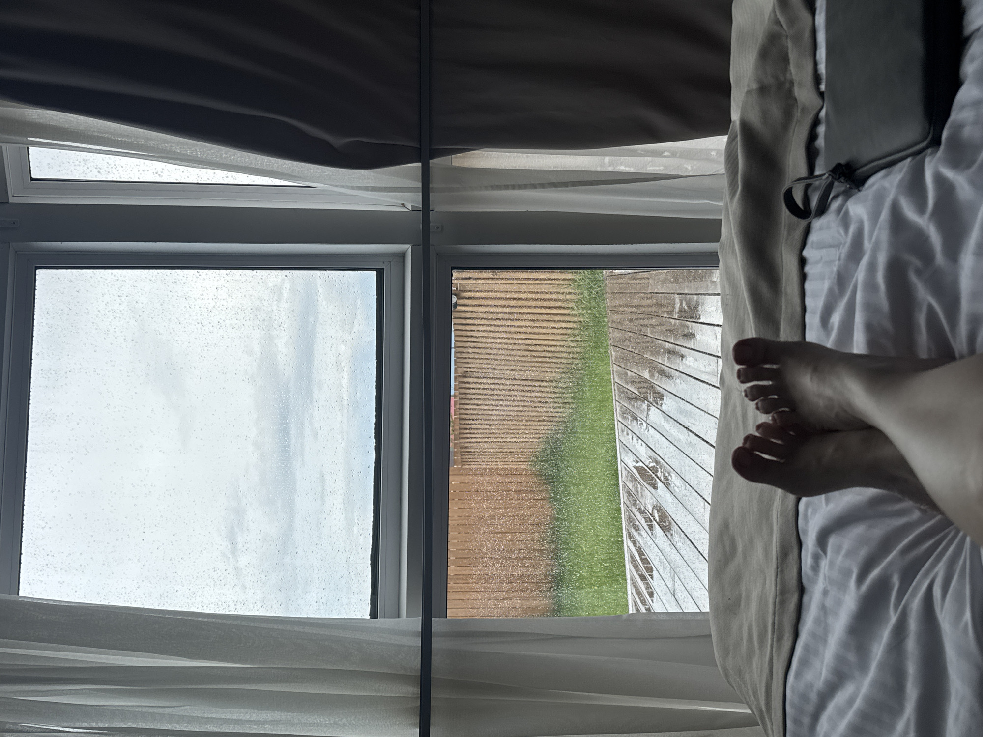 The image depicts a view from a window, likely in a bedroom, looking out onto a rainy day. The window is divided into two panes. The left pane shows a sky with visible raindrops, suggesting it's raining heavily. The right pane looks out onto a wooden deck or patio area, also wet from the rain, with a strip of green grass beyond it.
In the foreground, on the right side of the image, a person's bare feet are visible, resting on white bedding. The feet are positioned crossed, with toes slightly curled. The bedding appears soft and comfortable.
The overall scene evokes a sense of quiet relaxation, perhaps a cozy morning spent indoors due to the inclement weather. The focus is on the contrast between the wet, outdoor environment and the warm, dry interior. The time of day is not explicitly clear but the overcast sky suggests it could be daytime, possibly morning or afternoon. The location cues are minimal, but the description specifies it is Bolshoye Goloustnoye, Russia. There is no visible text in the image.