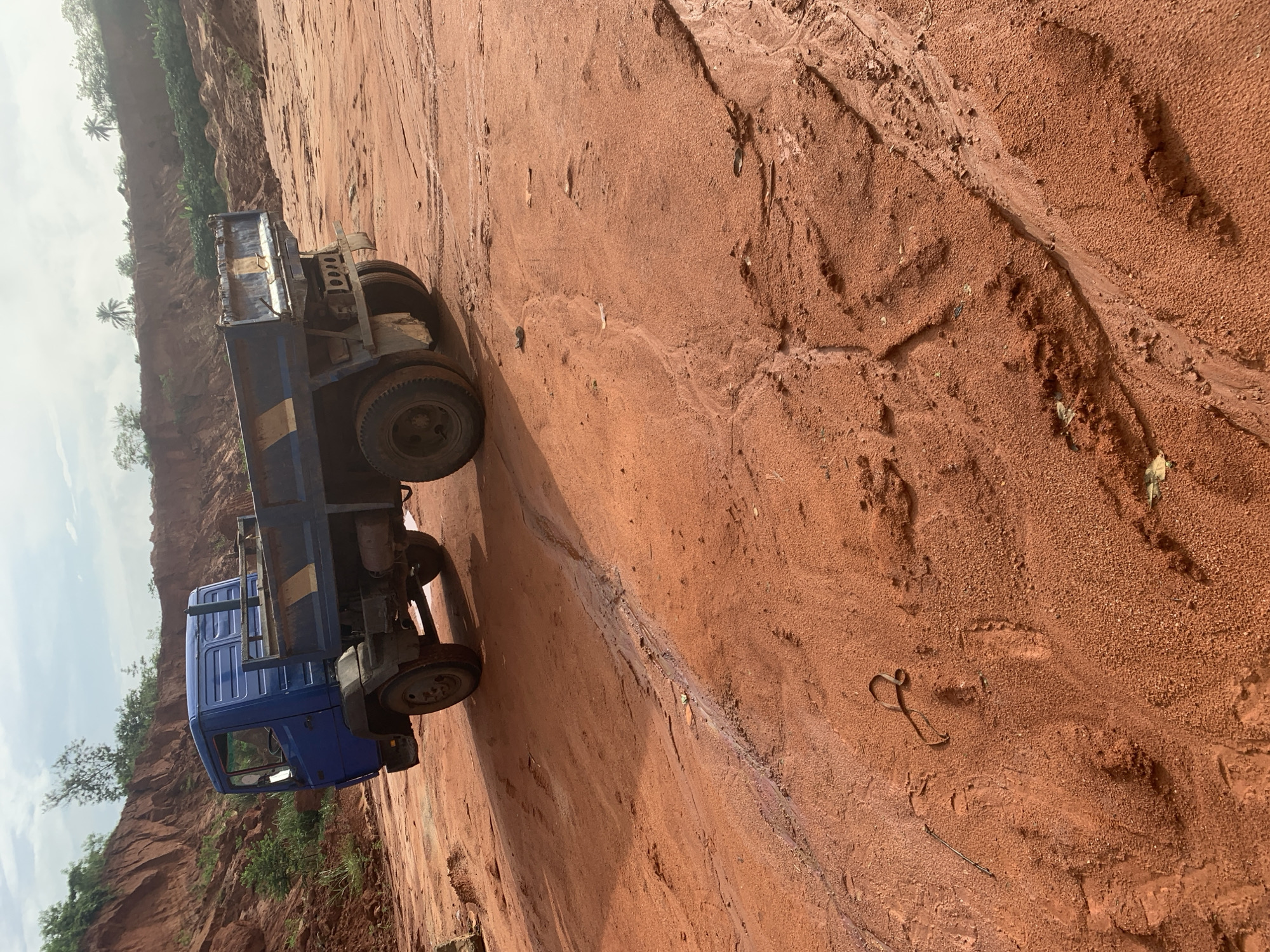 A blue dump truck is parked on a red dirt surface in what appears to be a quarry or construction site. The truck has a flatbed and dual rear wheels. The surrounding environment consists of reddish-brown soil banks and sparse vegetation. The sky is partly cloudy. The scene suggests an active or recently used industrial area, possibly in or around Ijesha-Ijebu, Nigeria.