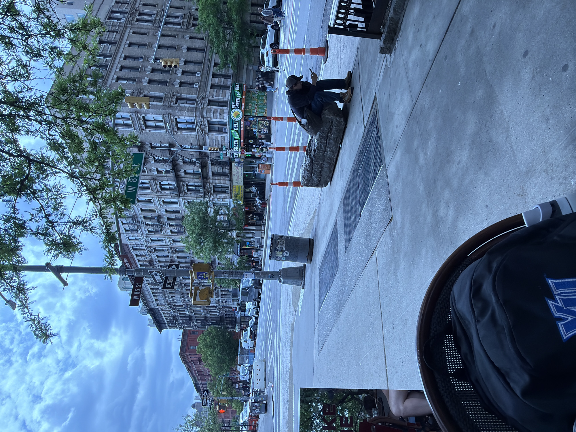 The image shows a street scene in New York City. The perspective is from an elevated position, likely from an outdoor seating area of a restaurant. A man is seated on a rock or concrete block on the sidewalk, looking at his mobile phone. He is wearing a dark jacket, jeans, and a baseball cap. Several traffic cones are lined up along the curb. Behind the man, across the street, is the storefront of "NEW GREEN Leaf DELI." Street signs indicating "W 85 St," "Amsterdam," and "W 8f" are visible on a lamp post. The buildings across the street are tall, older structures with detailed facades. The sky is cloudy. The environment appears to be urban and busy.