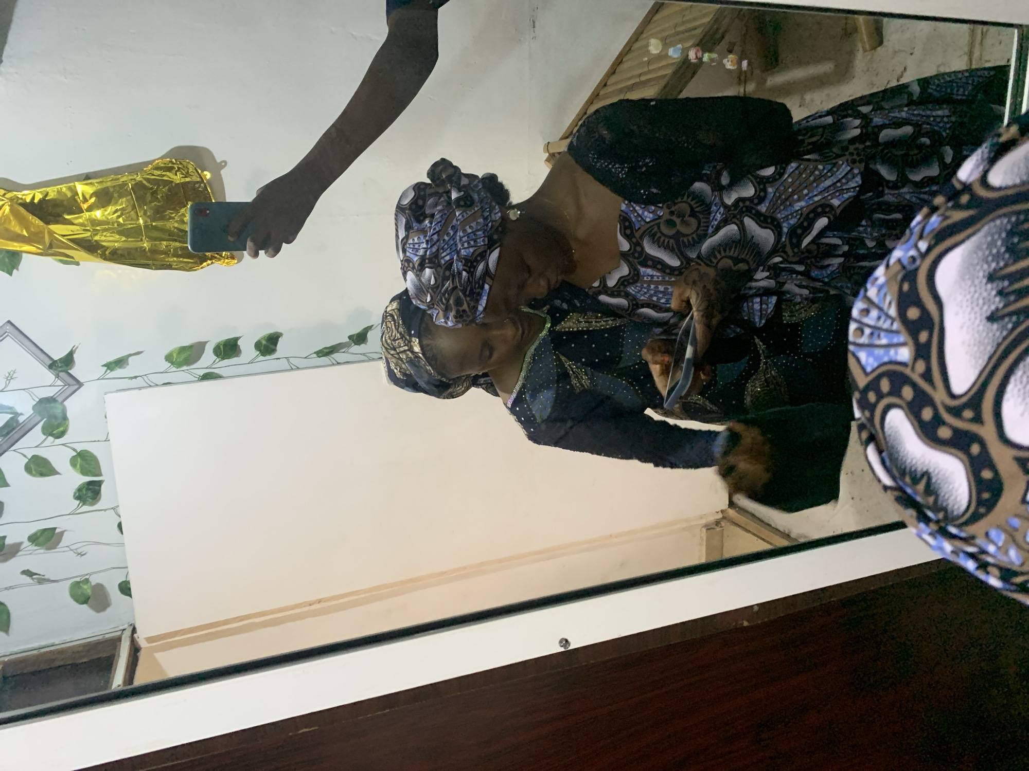 The image is a reflection in a mirror of a person taking a photo. Two women are visible in the mirror, both wearing head wraps and patterned clothing. One woman appears to be adjusting the other's head wrap or clothing. A hand holding a smartphone is visible, presumably taking the photo. A gold balloon is visible in the upper left corner of the mirror. The setting appears to be indoors, with a wall decorated with artificial vines. Jos, Nigeria is not evident within the image.