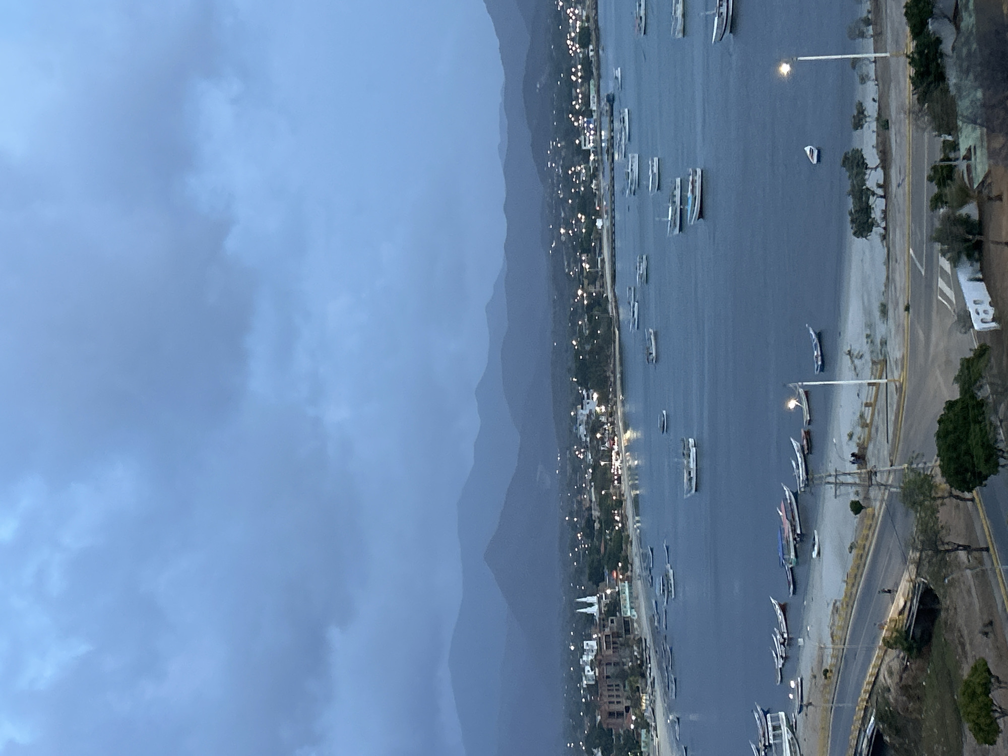 An elevated, wide-angle view captures Juan Griego Bay, Venezuela, at dusk or early night, characterized by a predominantly blue-grey and dark blue color palette under an overcast sky. A range of dark, undulating mountains forms the distant background. The midground features the illuminated town of Juan Griego curving along the bay, with numerous warm-toned city lights dotting the landscape. Prominently visible are the twin white steeples of a church, situated among other buildings on the left side of the bay. The dark blue water of the bay is populated by a multitude of anchored boats of varying sizes, predominantly white or light-colored, with some displaying colored tarps. Many smaller boats are beached or pulled up onto the sandy shore in the left-midground.
The foreground is dominated by a two-lane road or highway, featuring yellow and white lane markings, that parallels the coastline. A bridge structure is visible on the far left of the road. Two bright, warm-toned streetlights are operational, one on each side of the lower frame, casting light onto the road and immediate surroundings. Indistinct figures are discernible on the road and gathered on the sandy area near the right streetlight. Scattered trees and bushes line the roadside and parts of the sloped terrain leading down to the bay.