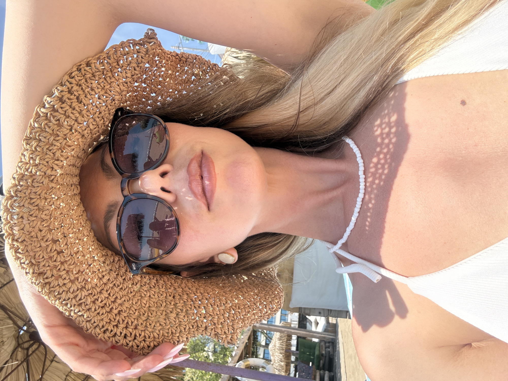 A fair-skinned individual is depicted in a close-up, chest-up selfie format, facing the camera under bright sunlight in Nesebar, Bulgaria. The person wears a wide-brimmed, light brown straw hat with a woven texture, dark tortoiseshell sunglasses, a white beaded choker, and a white ribbed bikini top. The right hand (viewer's left), with long manicured nails, grasps the hat brim, while the left arm (viewer's right) is raised with the elbow visible, positioned behind the hat. Long blonde hair falls over the shoulders. A small, circular, light-colored earring is visible on the person's left ear. The sunglasses reflect a beach scene, including a person taking a photograph, another individual, a thatched umbrella, and sand. The background, partially obscured by the hat and person, reveals a beach club environment with a purple-painted wooden pillar, a white lifebuoy, green outdoor seating, and a building in the distance on the left side, with a clear blue sky visible on the right. Strong shadows are cast by the hat on the person's chest and by other elements in the background. A distinct mole is present on the upper right side of the person's chest.